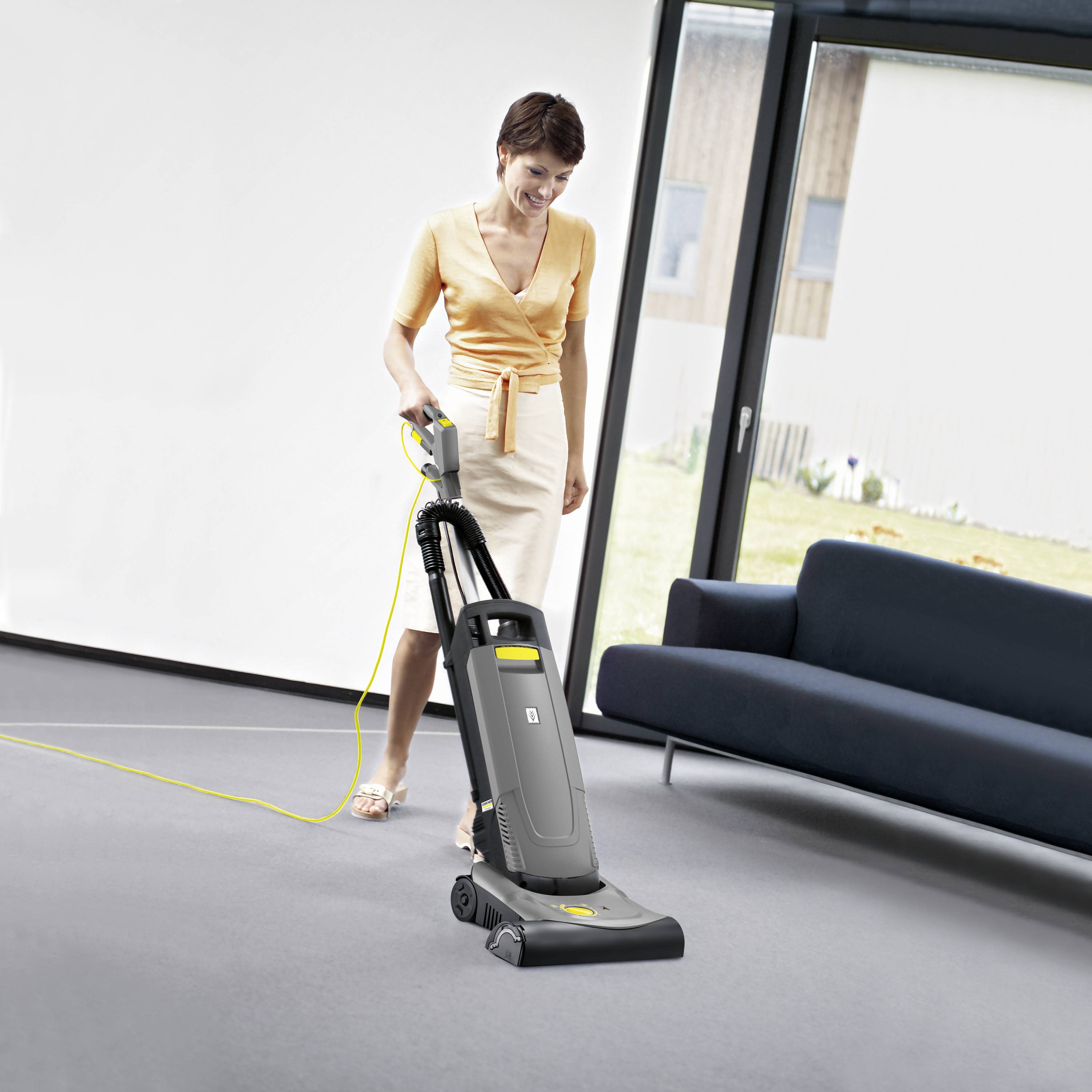 Une femme passe l'aspirateur sur le tapis dans un salon. Elle porte un chemisier orange et une jupe couleur crème.