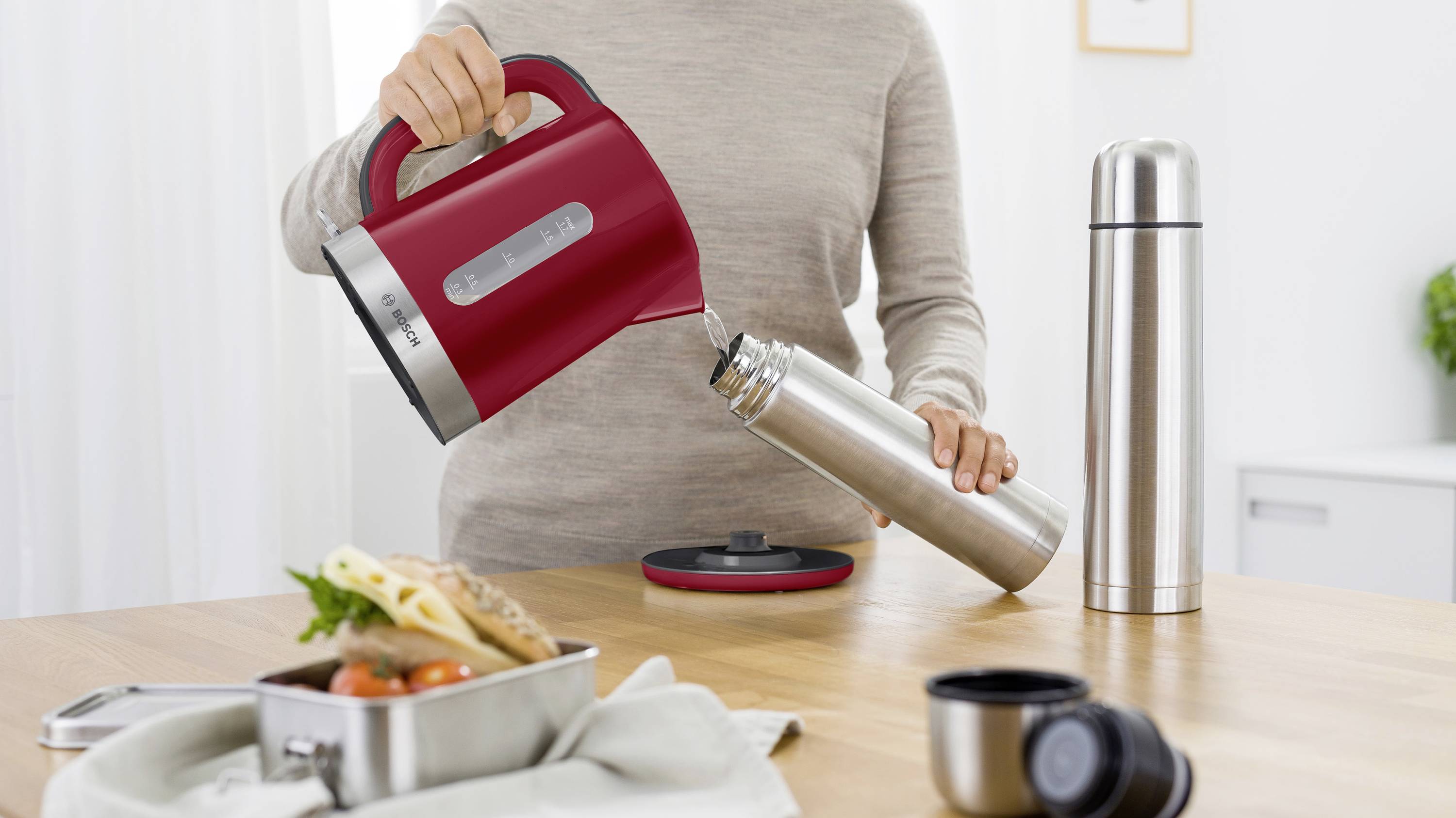 Une personne verse de l'eau chaude d'une bouilloire rouge dans un thermos argenté posé sur une table avec une boîte à pain et une tasse.