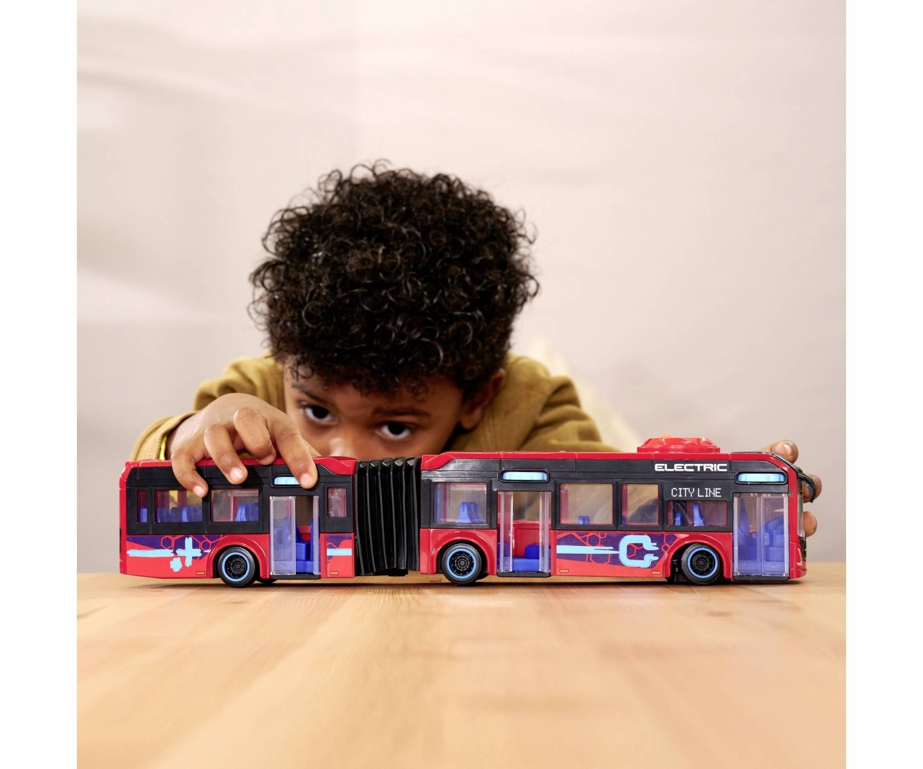 Un enfant joue avec un grand bus miniature sur une table. Le bus est rouge avec des détails bleus et blancs et porte l'inscription 'ELECTRIC'.