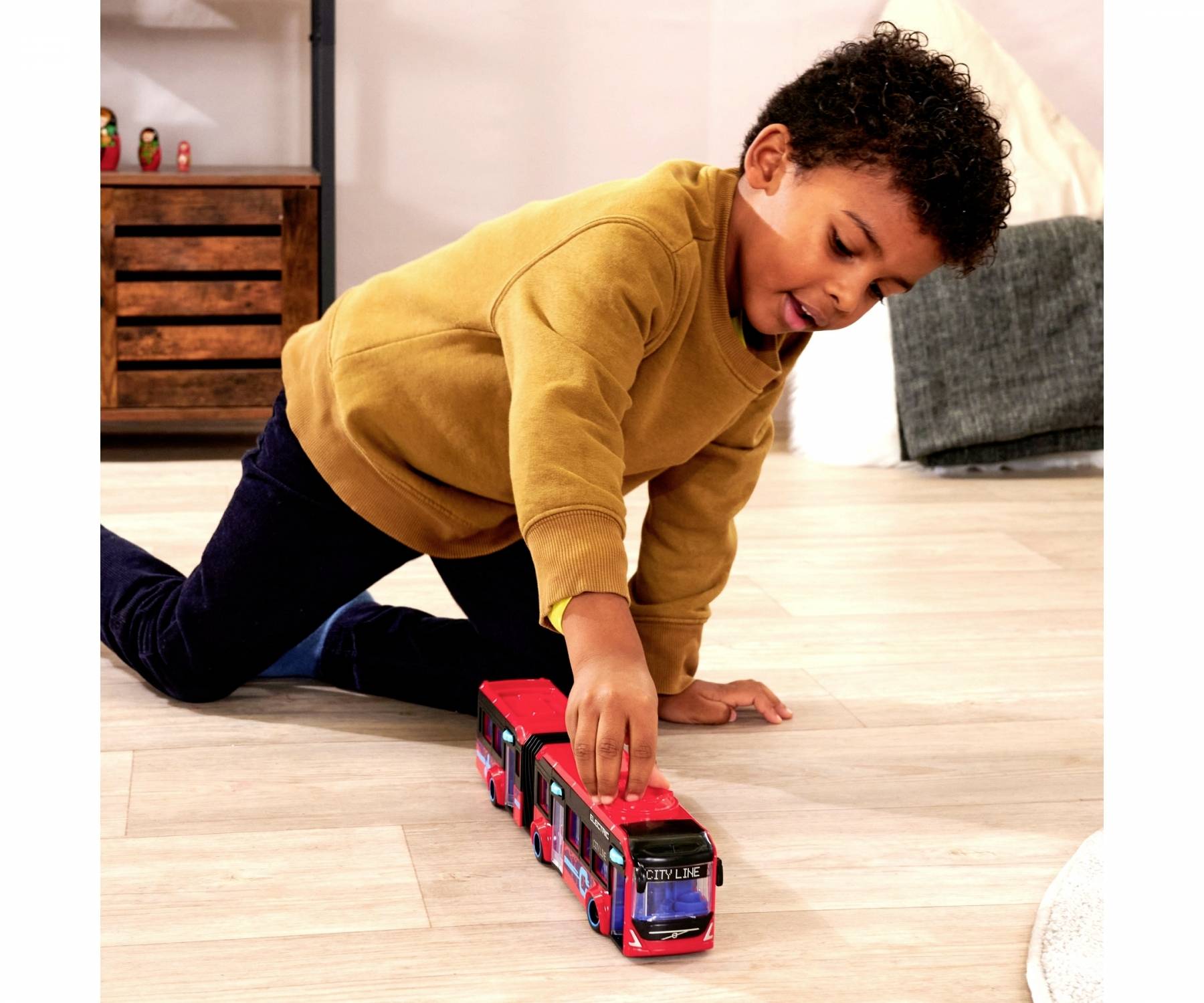 Un enfant joue avec un bus jouet rouge sur un sol en bois. Il porte un pull jaune et un jean bleu.