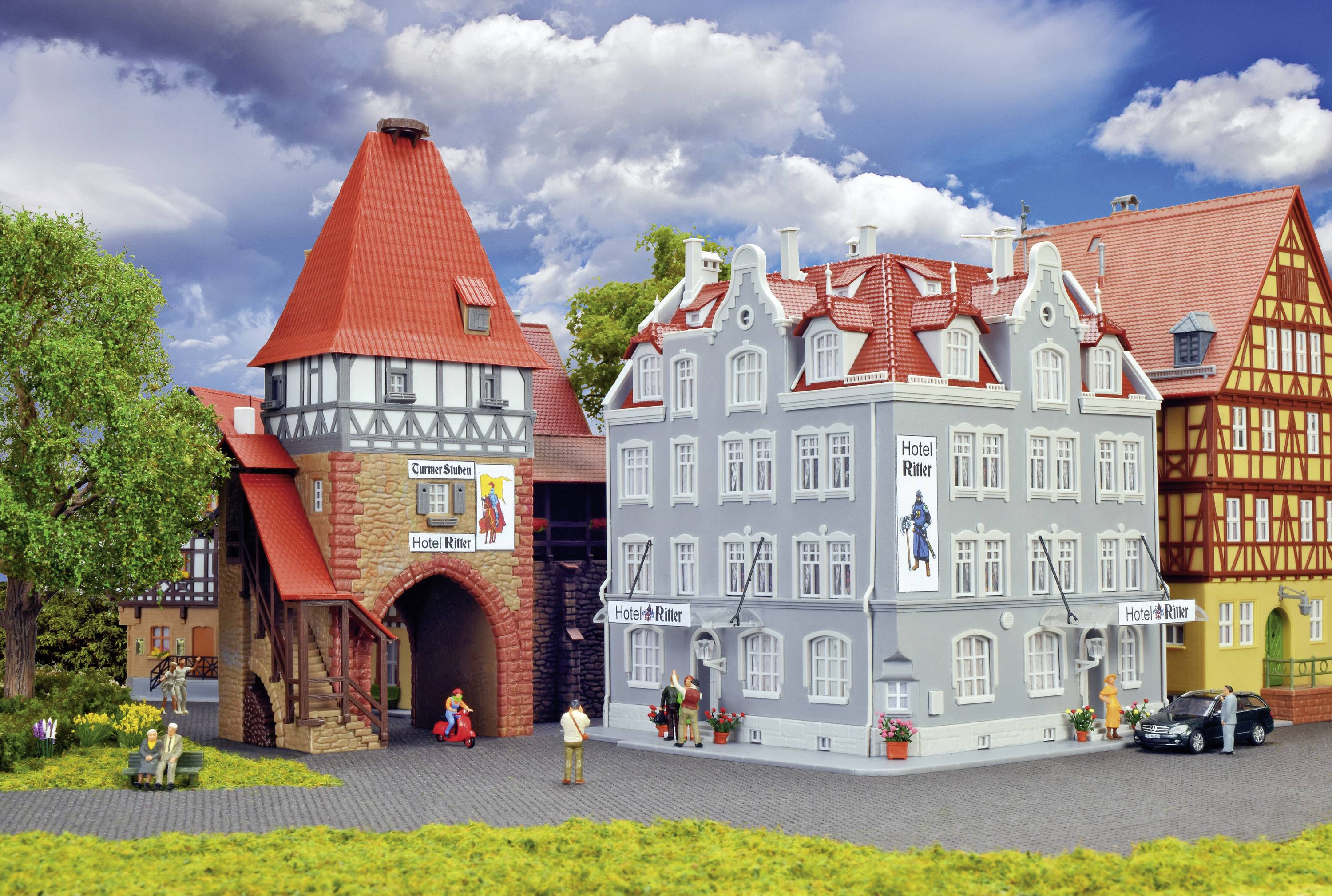 Un modèle miniature montre une rue avec un bâtiment à colombages, un hôtel et une tour. Des personnes et une voiture sont visibles.