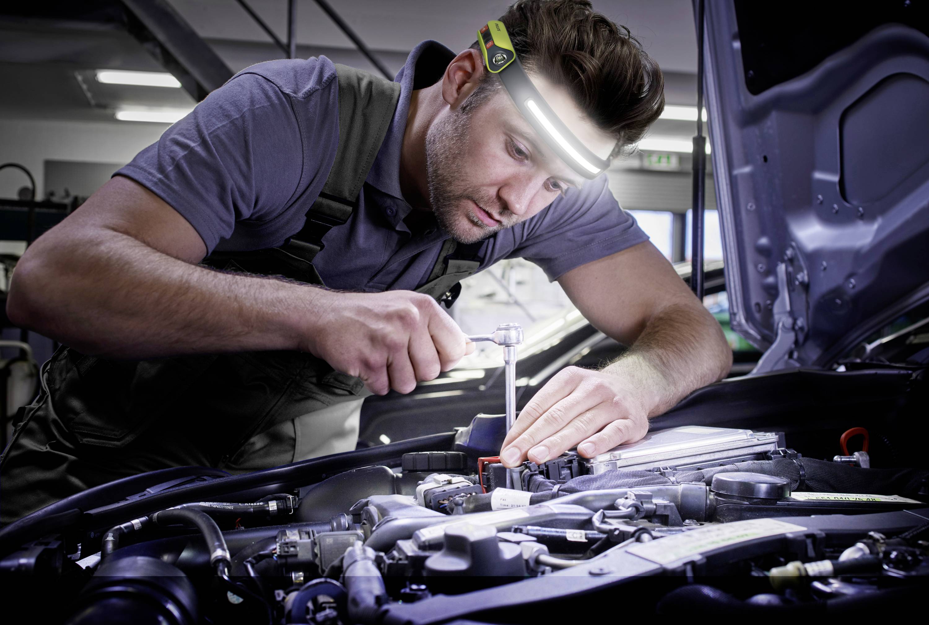Un mécanicien, concentré sur un moteur de voiture, porte une lampe frontale. À l'arrière-plan, des outils et l'éclairage de l'atelier sont visibles.