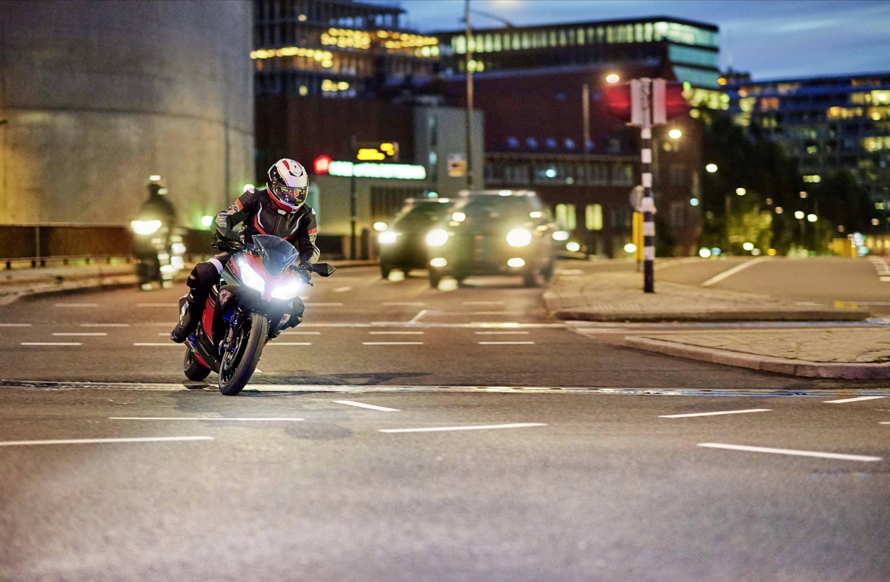 Un motocycliste traverse un carrefour urbain au crépuscule, entouré de voitures et de bâtiments éclairés en arrière-plan.