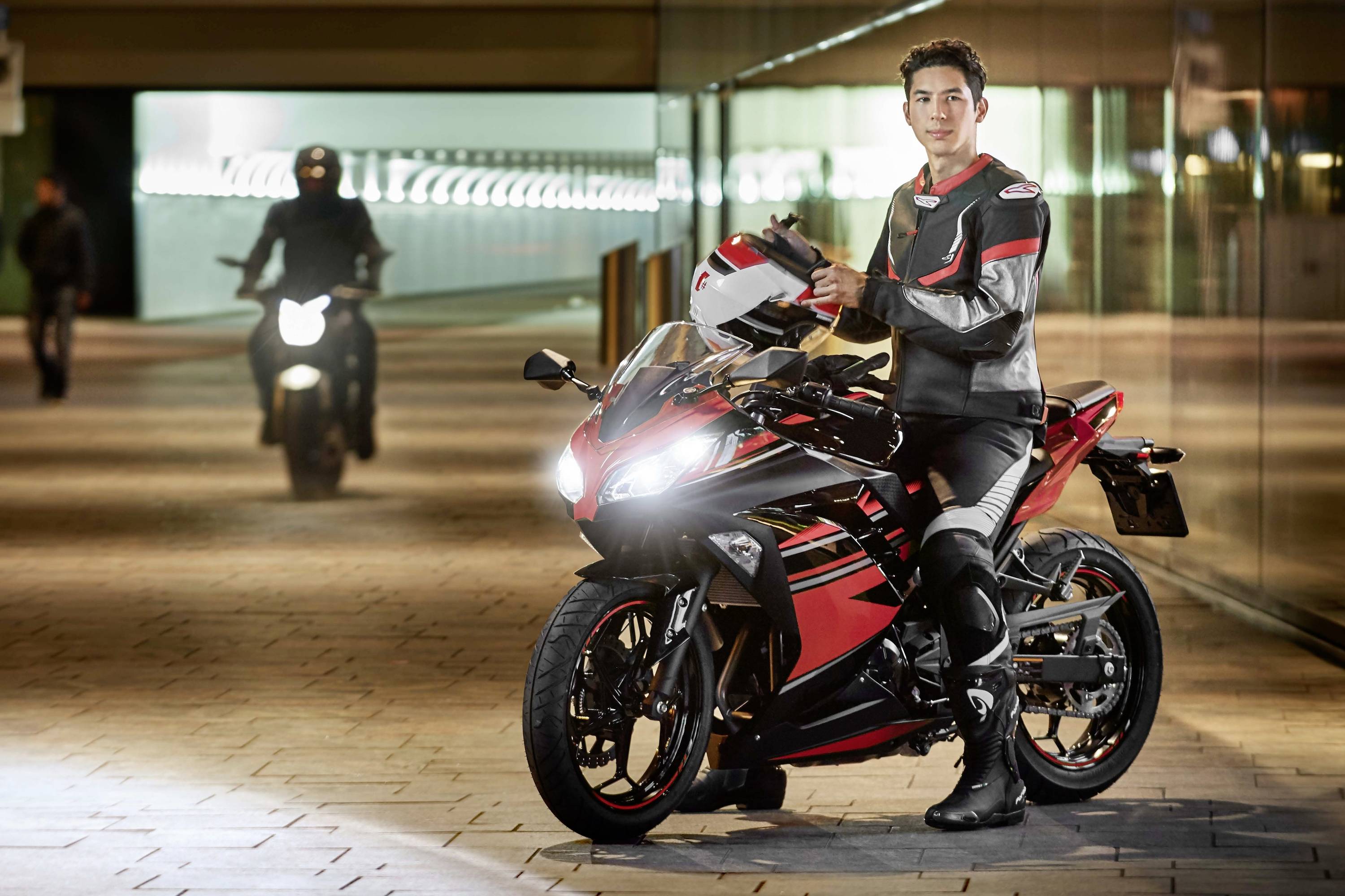 Un motard en tenue de protection tient un casque à côté d'une moto sportive rouge dans un passage éclairé. Une autre moto est visible en arrière-plan.