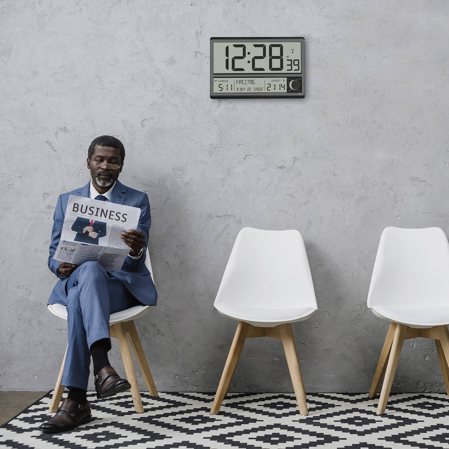Un homme en costume est assis sur une chaise et lit un journal dont le titre est 'Business'. Deux chaises vides sont placées à côté de lui. Une horloge indique 12h28.