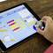 Une tablette affiche une application de programmation pour enfants. Une main déplace un petit robot à l'écran pour le guider.