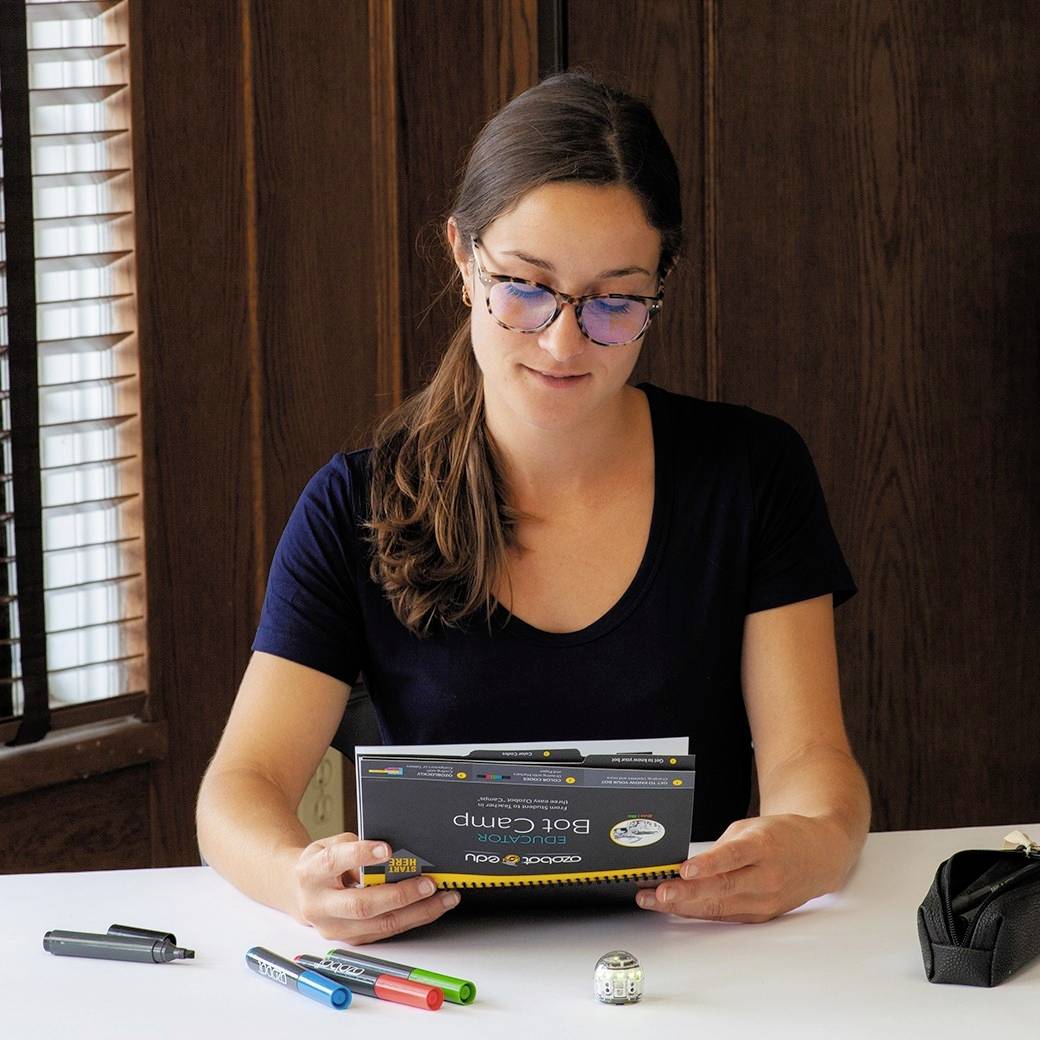 Une personne est assise à une table avec une tablette sur laquelle est écrit 'Bootcamp'. Des stylos et un dé sont posés devant elle. Elle semble travailler de manière concentrée.