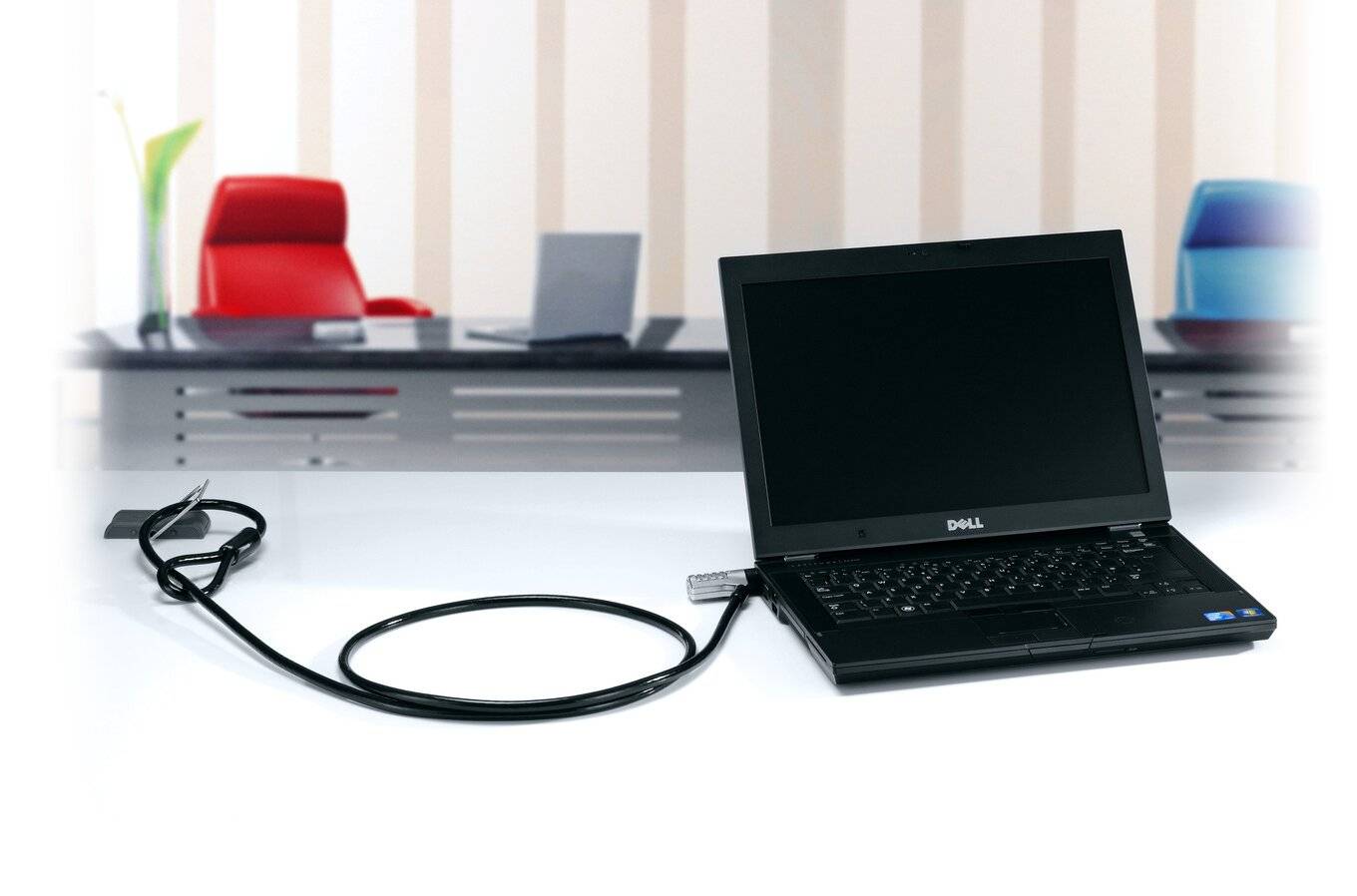 Un ordinateur portable ouvert, relié à un câble de sécurité. En arrière-plan, un espace de bureau moderne avec des chaises et des tables.