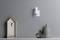Horloge en bois en forme de maison avec un cadran noir à gauche, trois vases à droite sur une étagère devant un mur gris avec un interrupteur blanc.