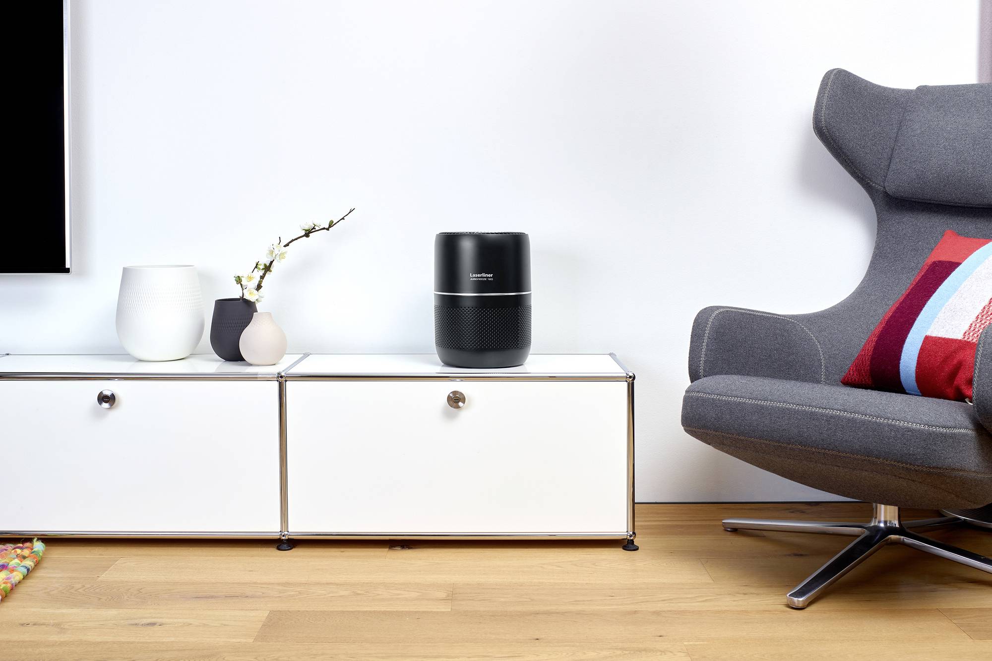 Un espace de vie moderne avec un haut-parleur noir sur un buffet blanc. À côté, des vases contenant une orchidée. À droite, un fauteuil gris avec un coussin coloré.