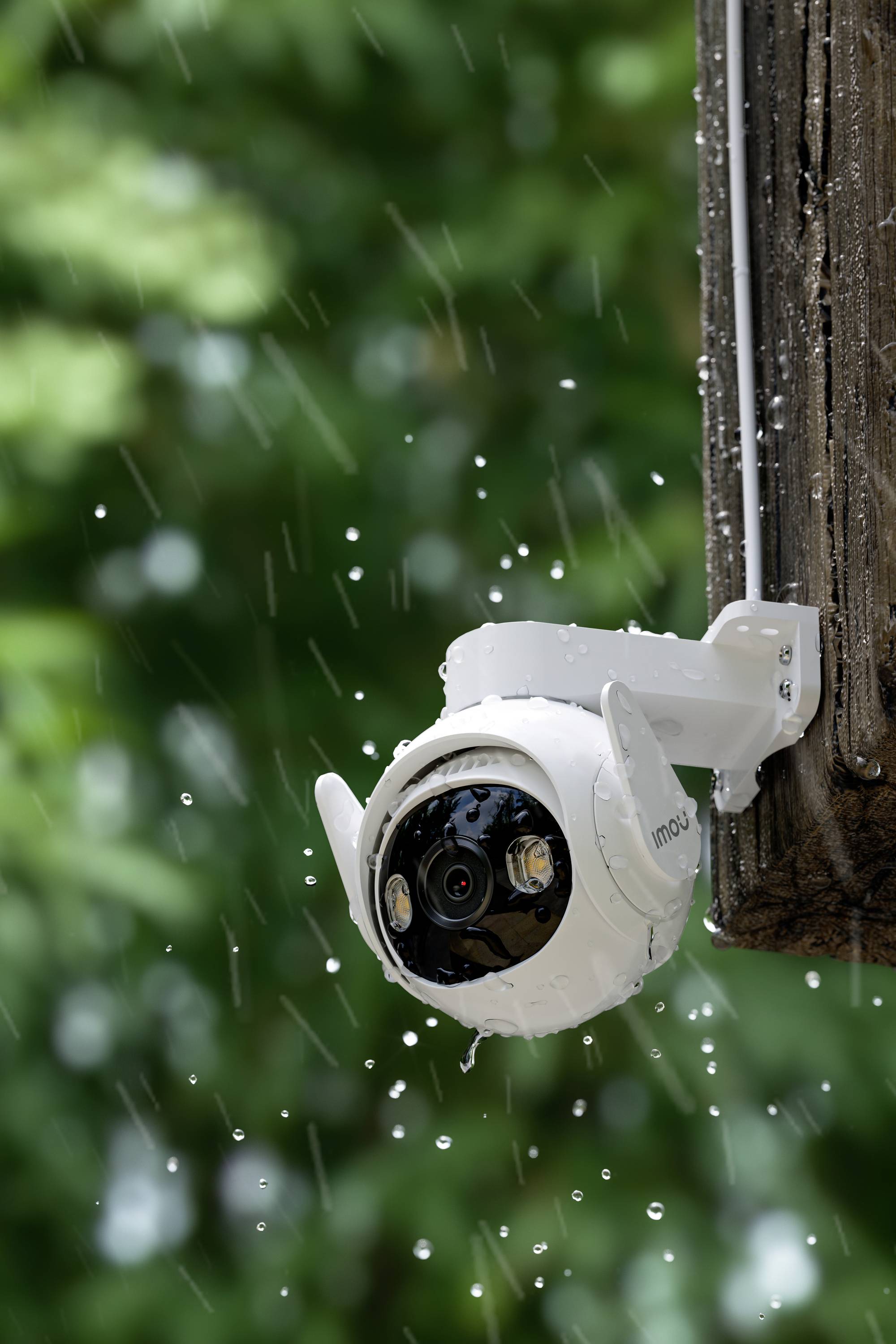 Caméra de surveillance sous la pluie, fixée sur un poteau en bois, avec un arrière-plan flou de feuilles vertes.