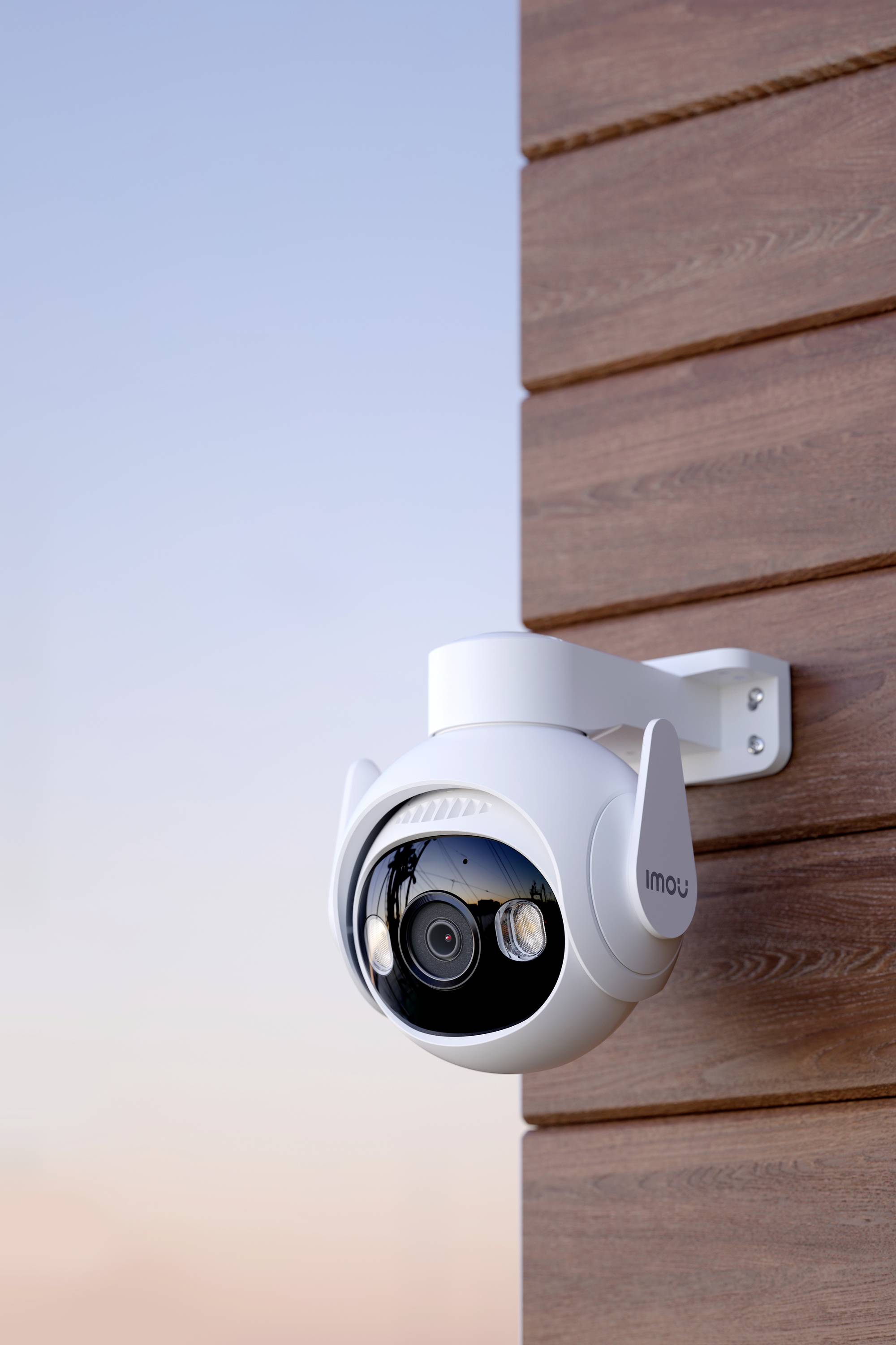 Une caméra de surveillance blanche est fixée à un mur en bois, dirigée vers le ciel en arrière-plan. Elle présente un design moderne et sphérique.