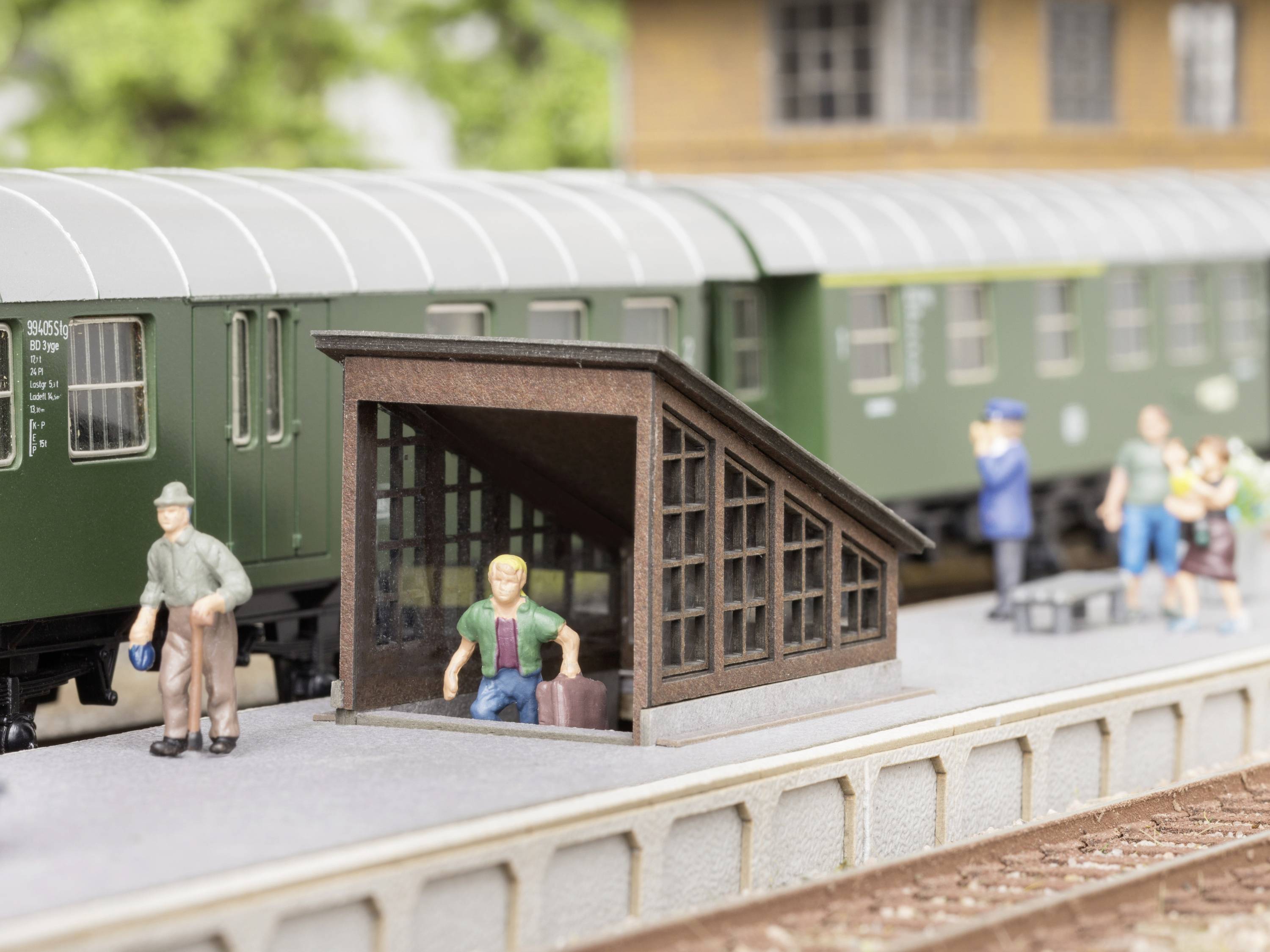 Scène en miniature : Des personnes sur un quai à côté d'un train vert, certaines portant des bagages. Un homme emprunte un escalier couvert.