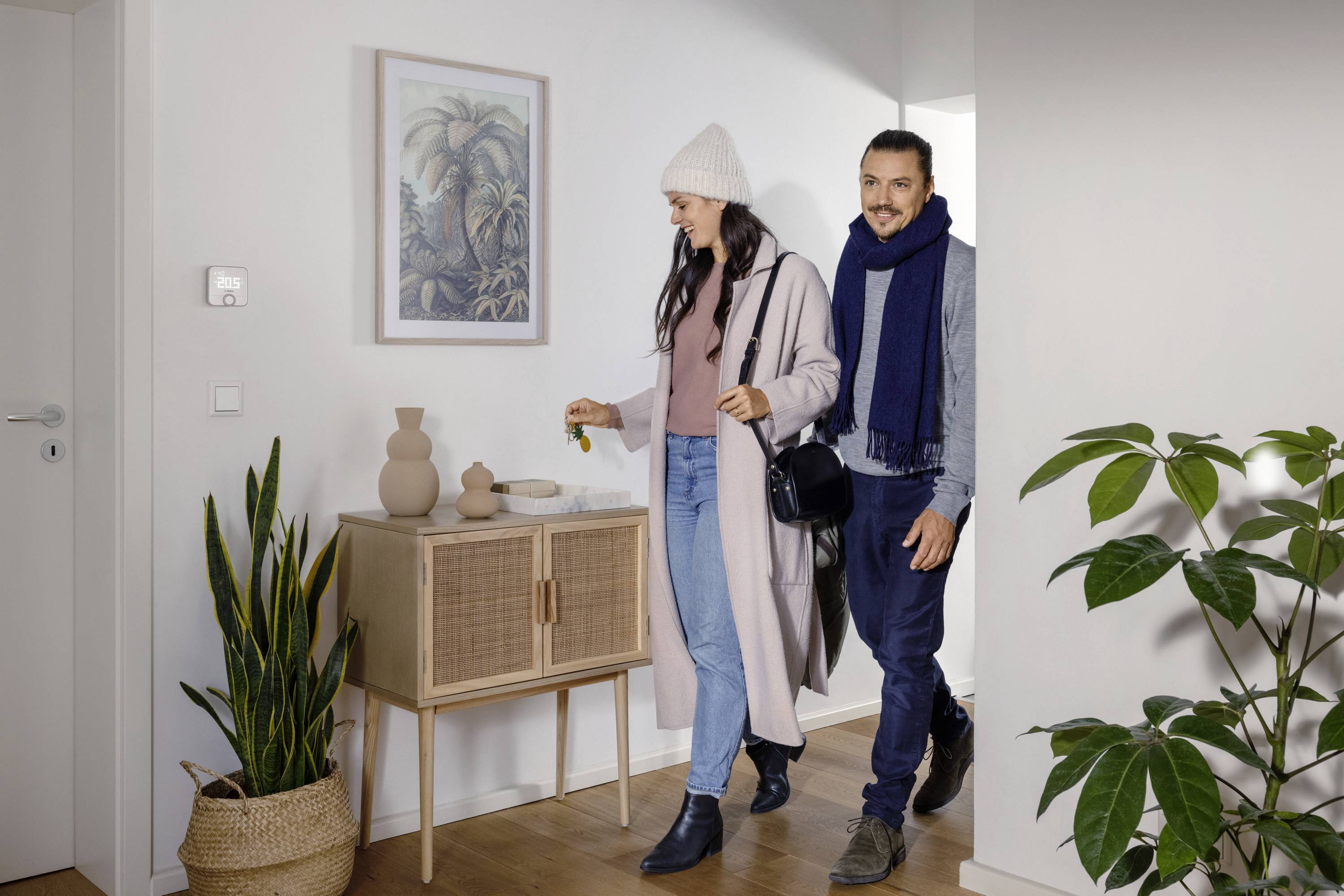 Un homme et une femme entrent dans une pièce en souriant, tous deux vêtus de vêtements d'hiver. Des plantes et un placard sont visibles en arrière-plan.