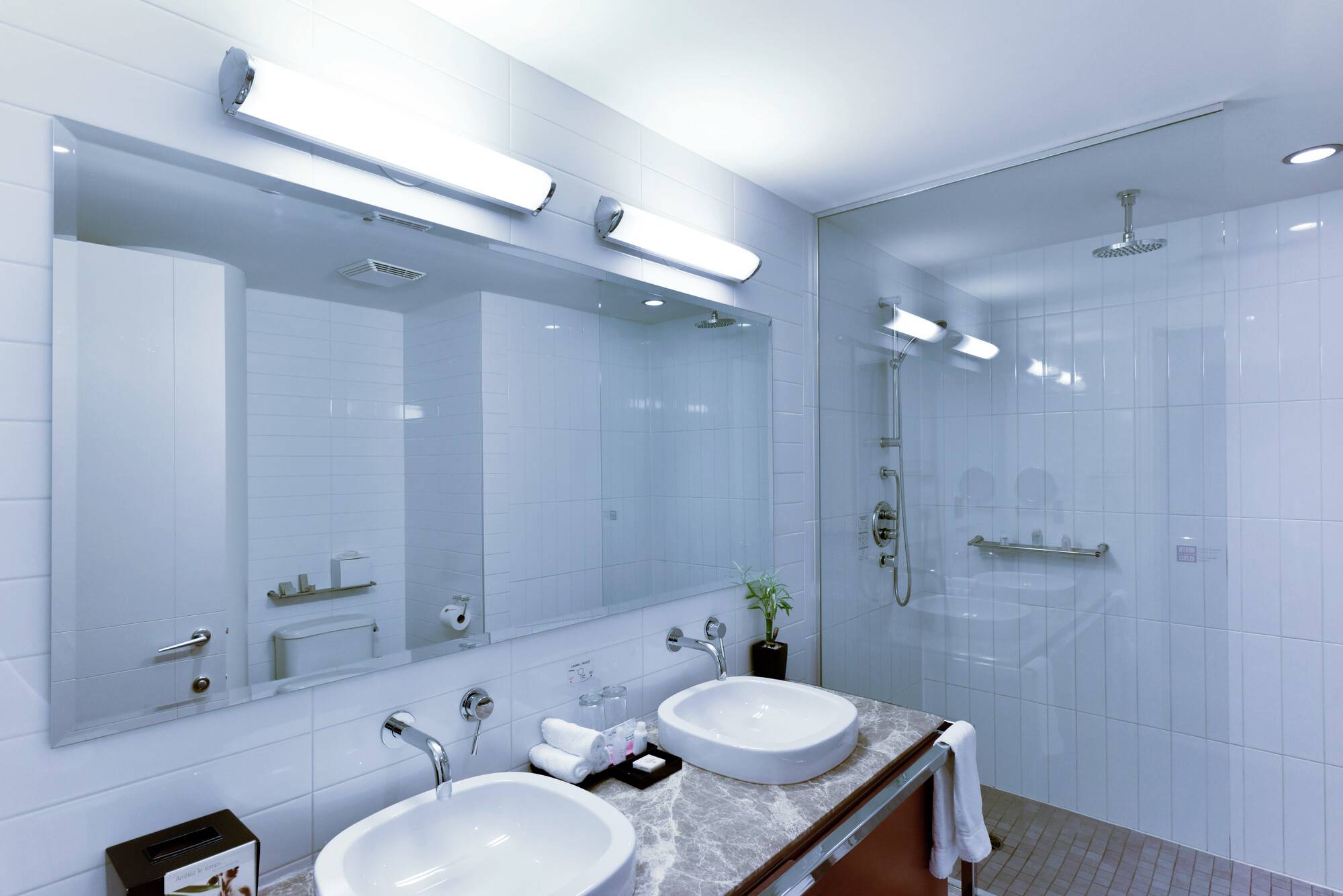 Salle de bain moderne avec deux lavabos, grand miroir et luminaires éclairés. Douche en arrière-plan, carrelage blanc sur les murs.