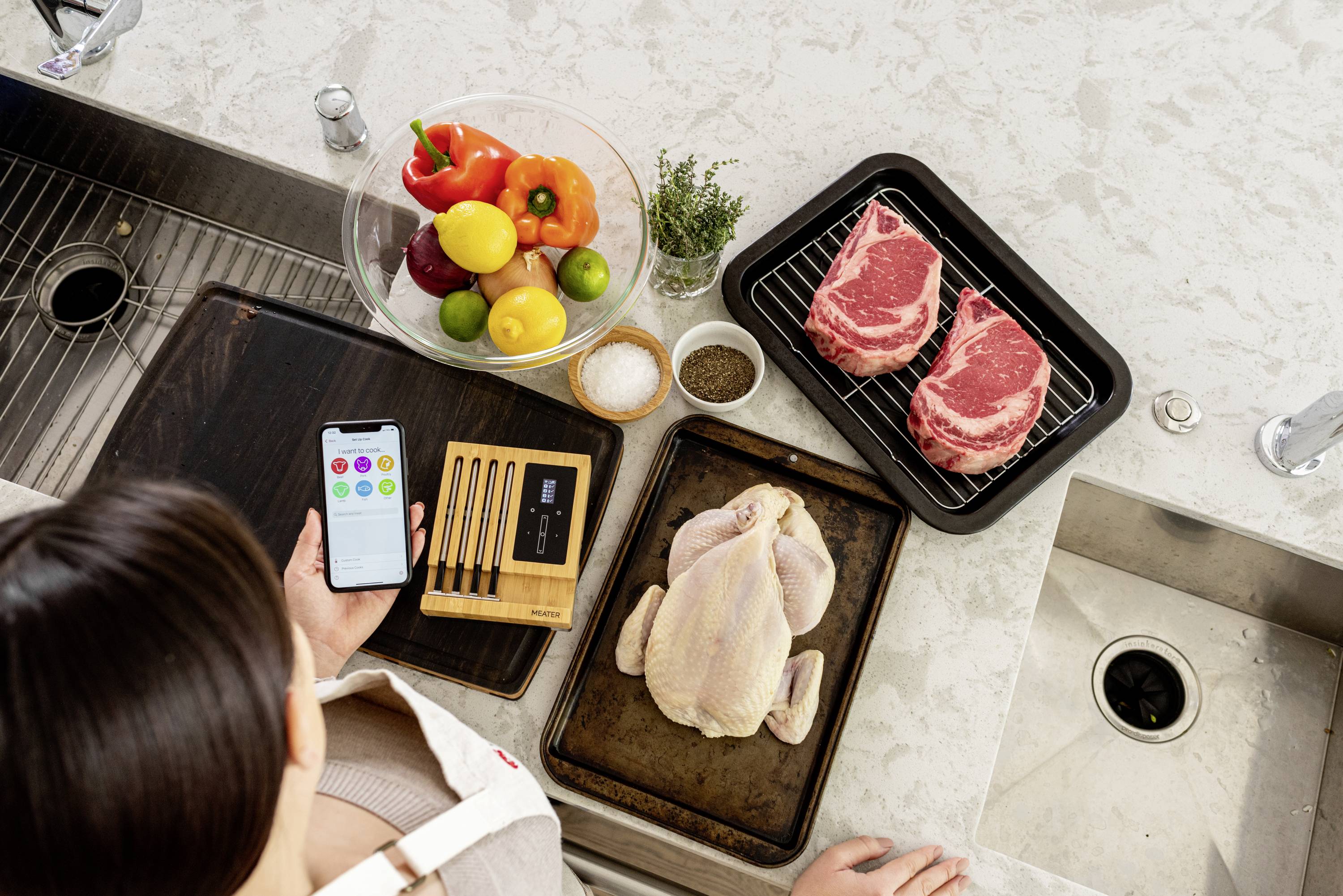 Une femme se tient dans la cuisine tenant un smartphone qui affiche une application de cuisine. Sur le plan de travail sont disposés des légumes, des steaks et un poulet.