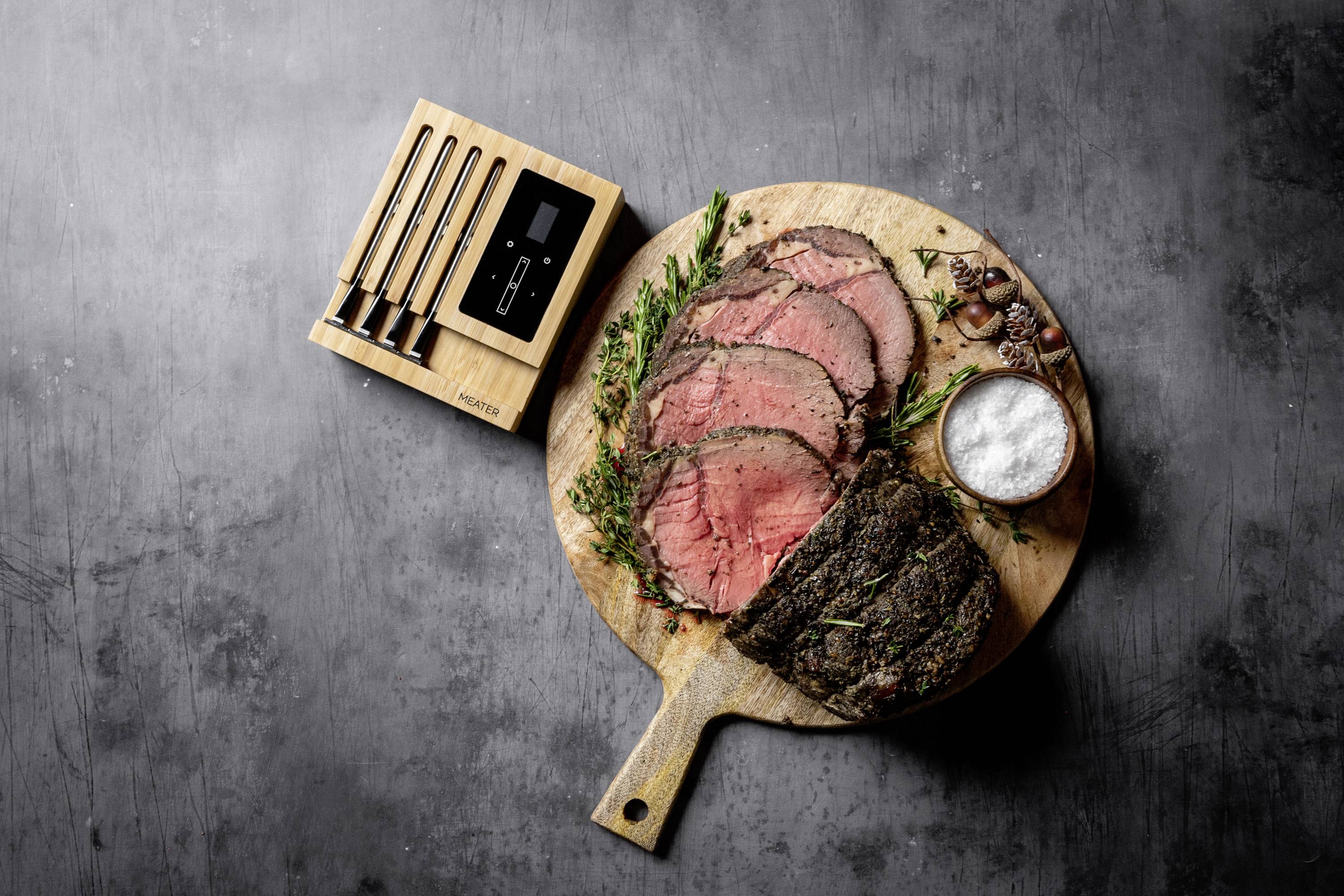 Une planche ronde en bois avec un rôti de bœuf tranché, des herbes fraîches et un bol de sel. À côté, un set de couteaux dans son support en bois.