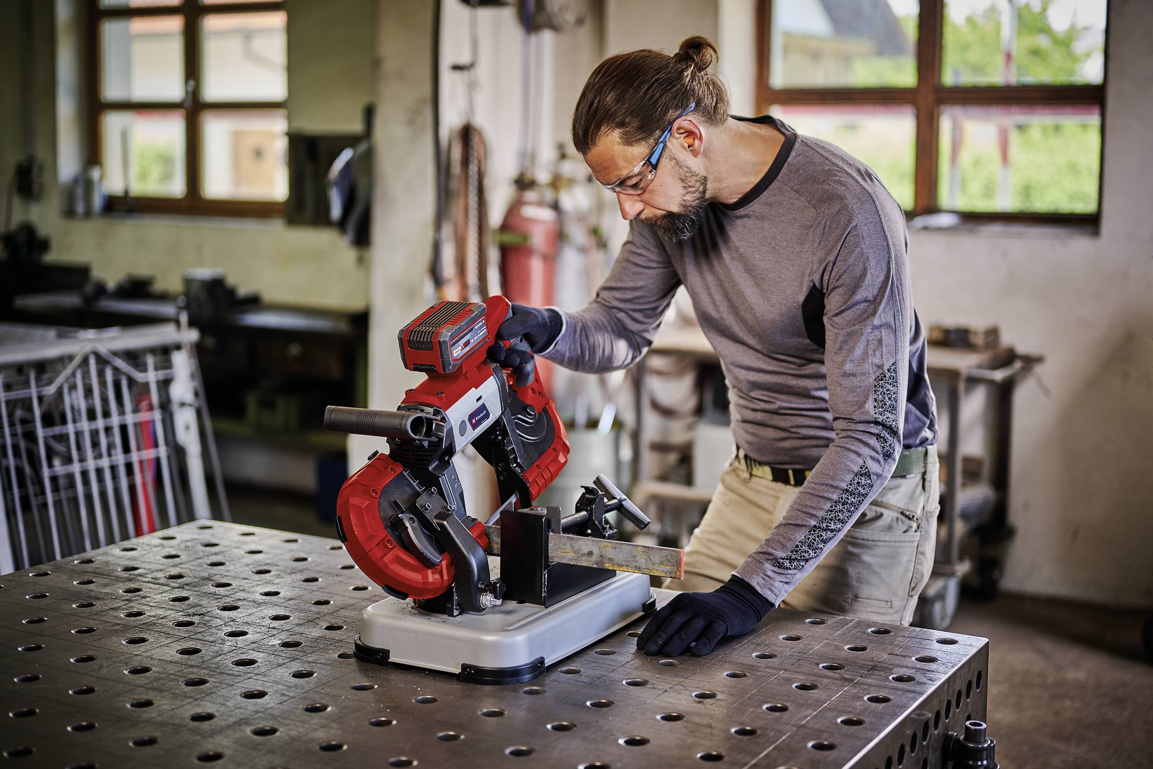 Un homme portant des lunettes de sécurité et des gants travaille avec concentration sur une scie à ruban métallique dans un atelier.