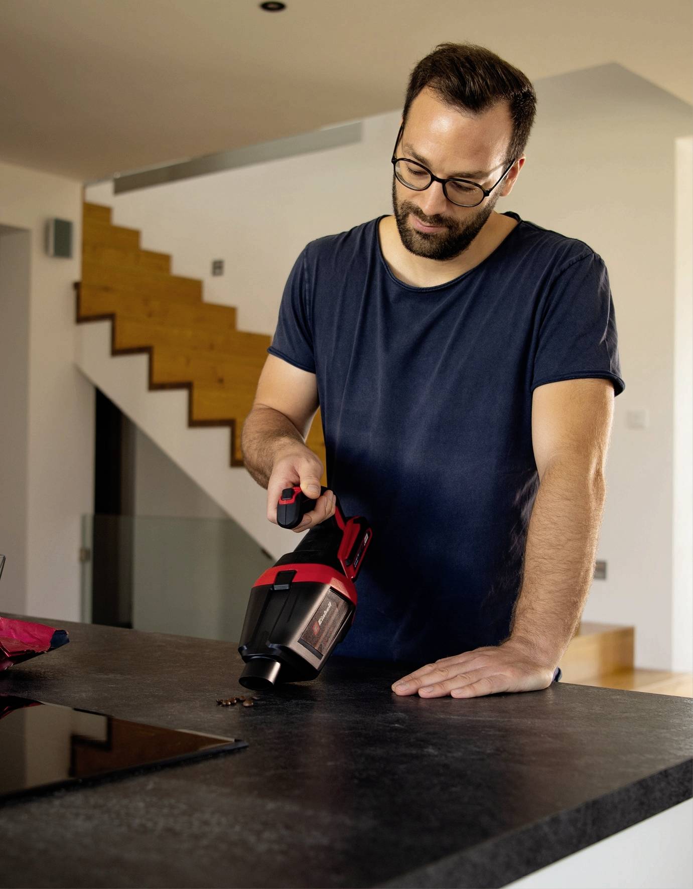 Un homme utilise un aspirateur à main pour enlever des miettes d'un plan de travail de cuisine. Des escaliers en bois sont visibles en arrière-plan.