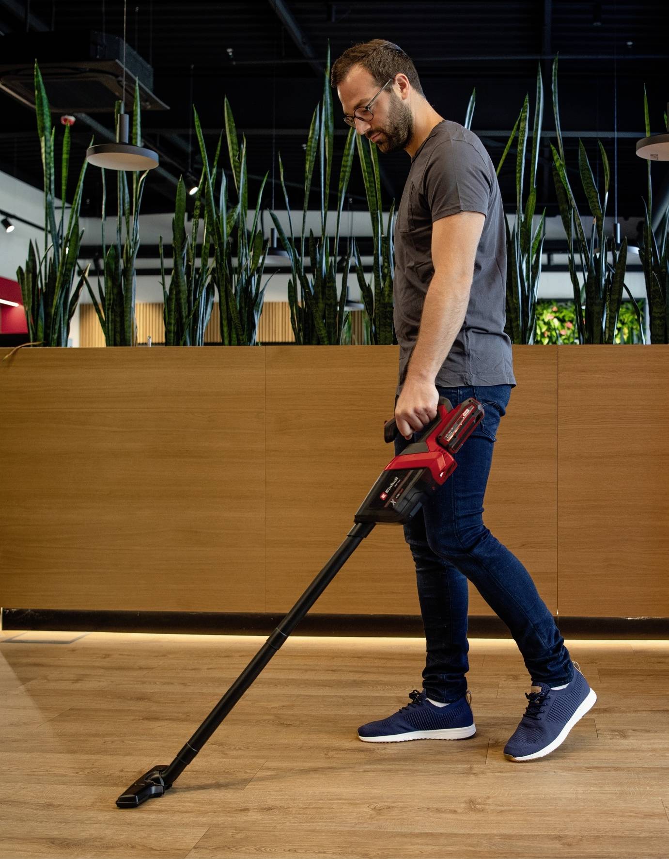 Une personne utilise un aspirateur sans fil pour nettoyer un parquet dans une pièce au design moderne avec des plantes en arrière-plan.