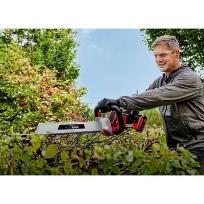 Un homme taille des buissons dans le jardin avec un taille-haie électrique. Il porte des lunettes de protection et des gants, et sourit avec concentration.