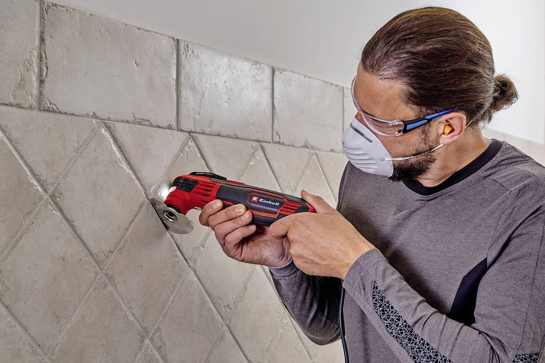 Un homme travaille avec un outil électrique sur un carreau d'un mur. Il porte des lunettes de protection et un masque.