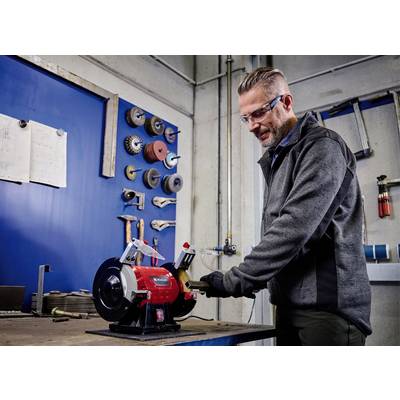 Un homme travaille un outil avec un appareil de meulage dans un atelier. Des outils et des meules sont accrochés au mur.