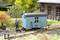 Un ouvrier en construction de maquettes, vêtu de bleu, se tient devant une petite caravane de chantier bleue dans un paysage ferroviaire avec un bâtiment en briques.