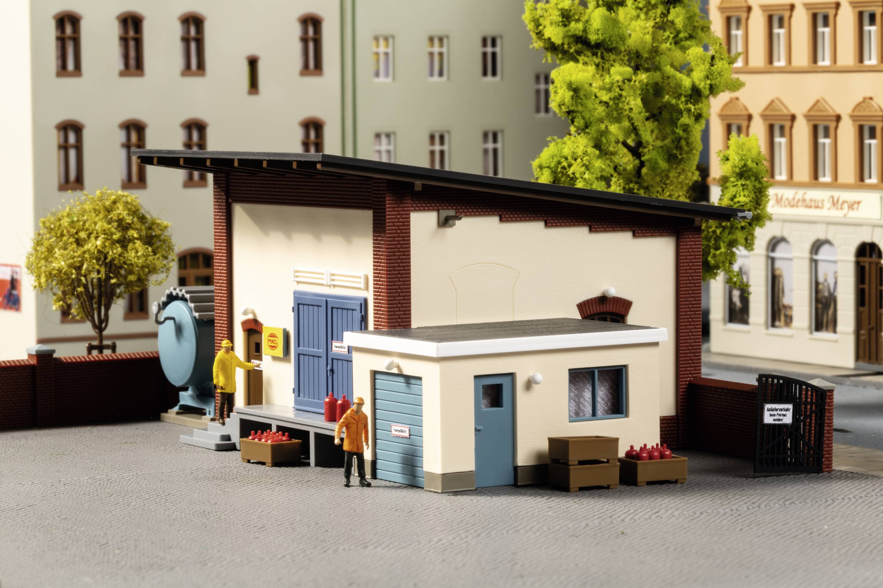 Une maquette de maison urbaine avec un quai de chargement, un arbre et des caisses au premier plan. D'autres maisons sont visibles à l'arrière-plan.