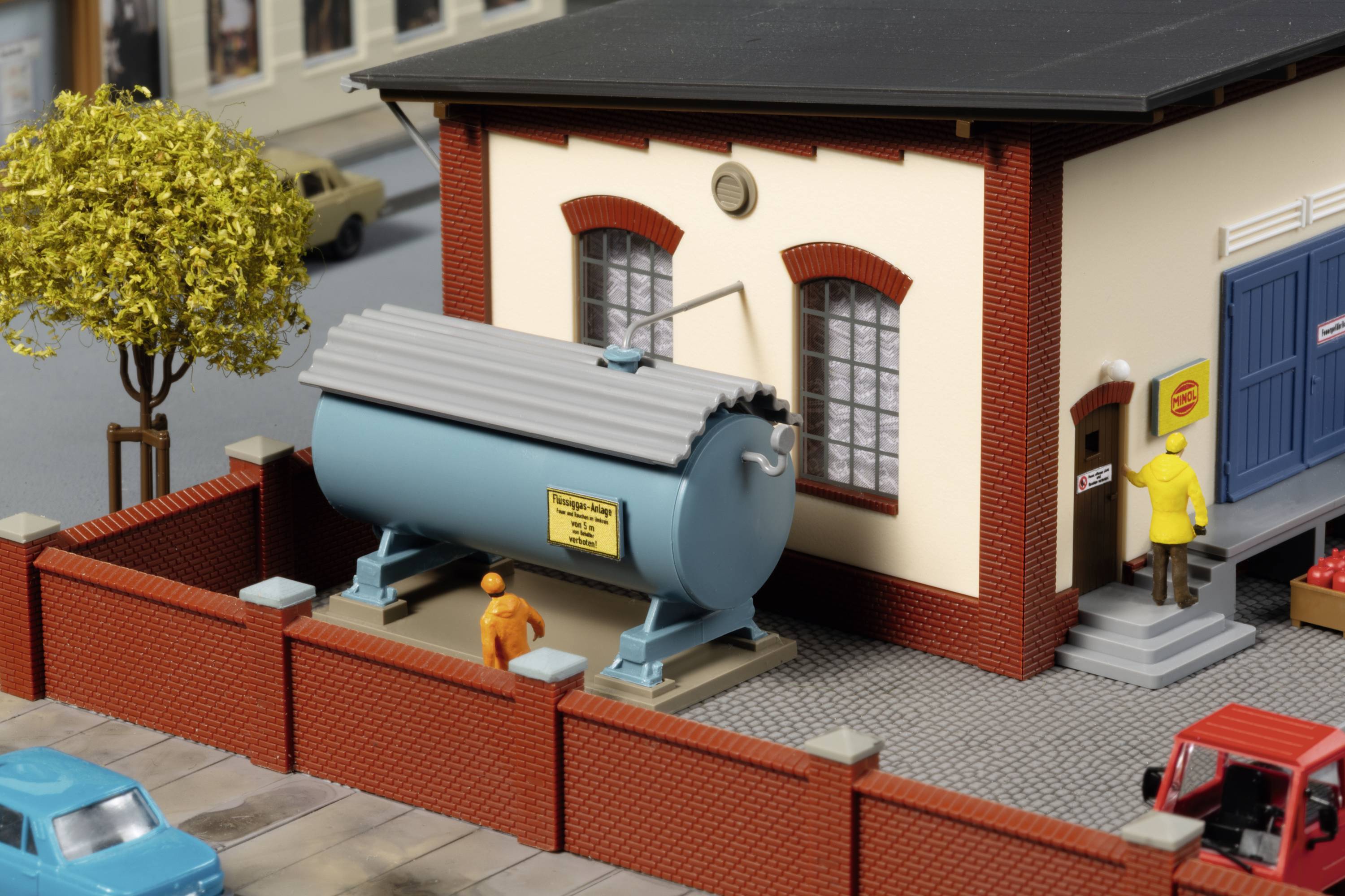 Un modèle miniature montre un bâtiment avec un réservoir bleu à côté, entouré d'un mur rouge. Deux personnages se tiennent à proximité.