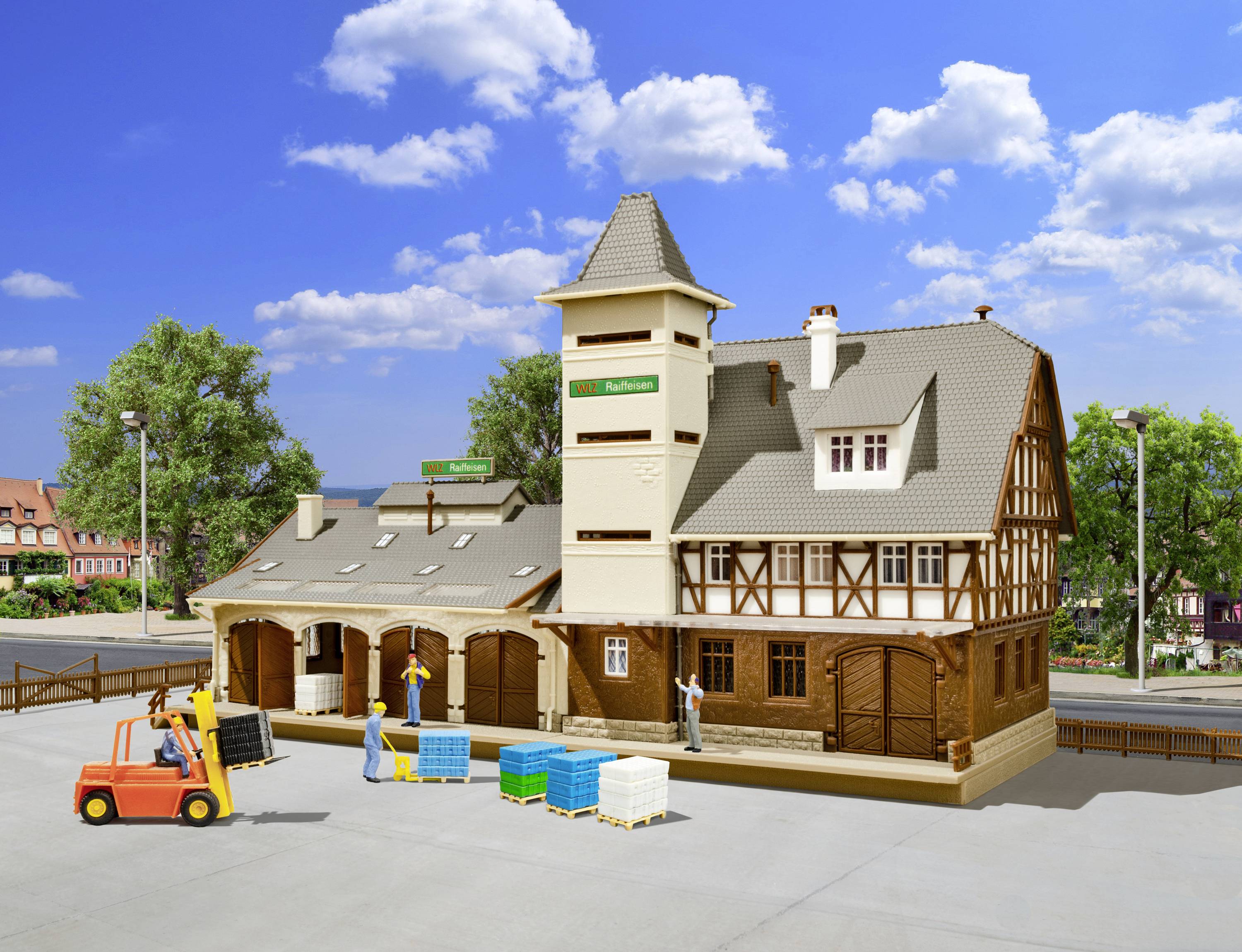 Maquette d'une gare avec un entrepôt de marchandises, un chariot élévateur et des figurines sur une surface bétonnée, entourée d'arbres et de ciel.