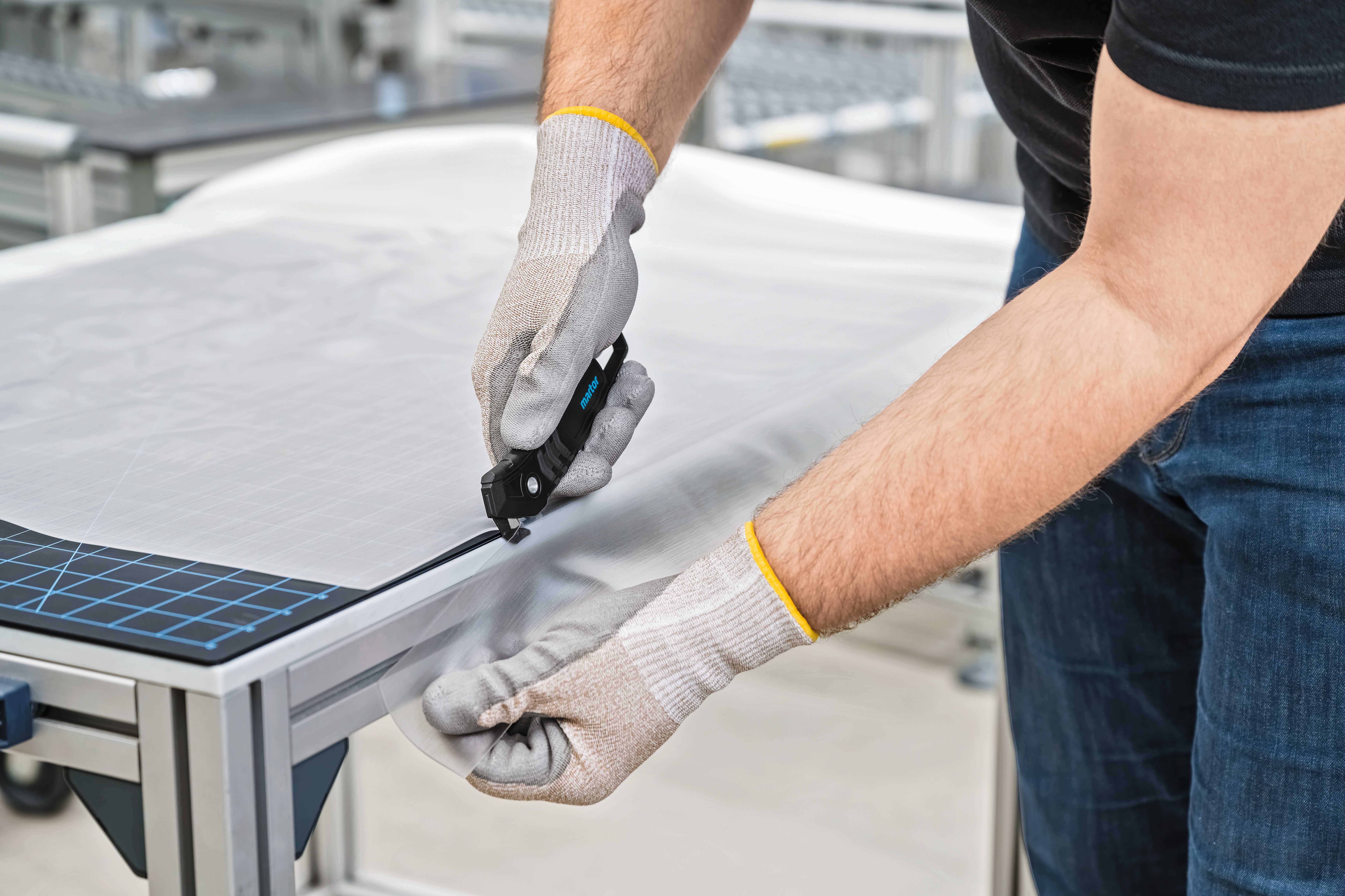 Une personne coupe soigneusement du tissu sur une table avec un couteau à découper, portant des gants de travail.