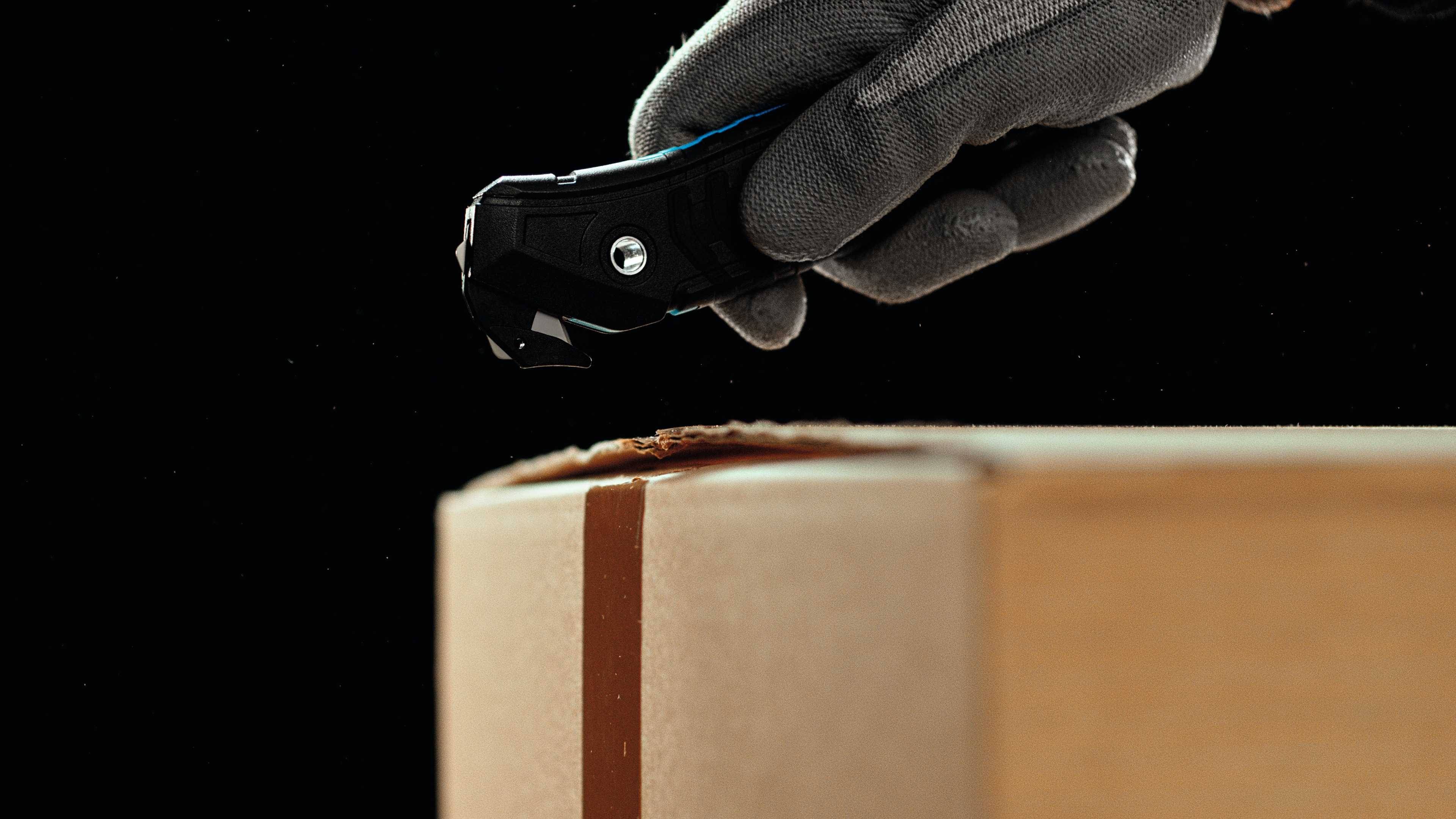 Une main gantée coupe un carton avec un couteau.