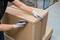 Une personne ouvre un grand carton brun avec un cutter dans un entrepôt. Elle porte des gants gris.