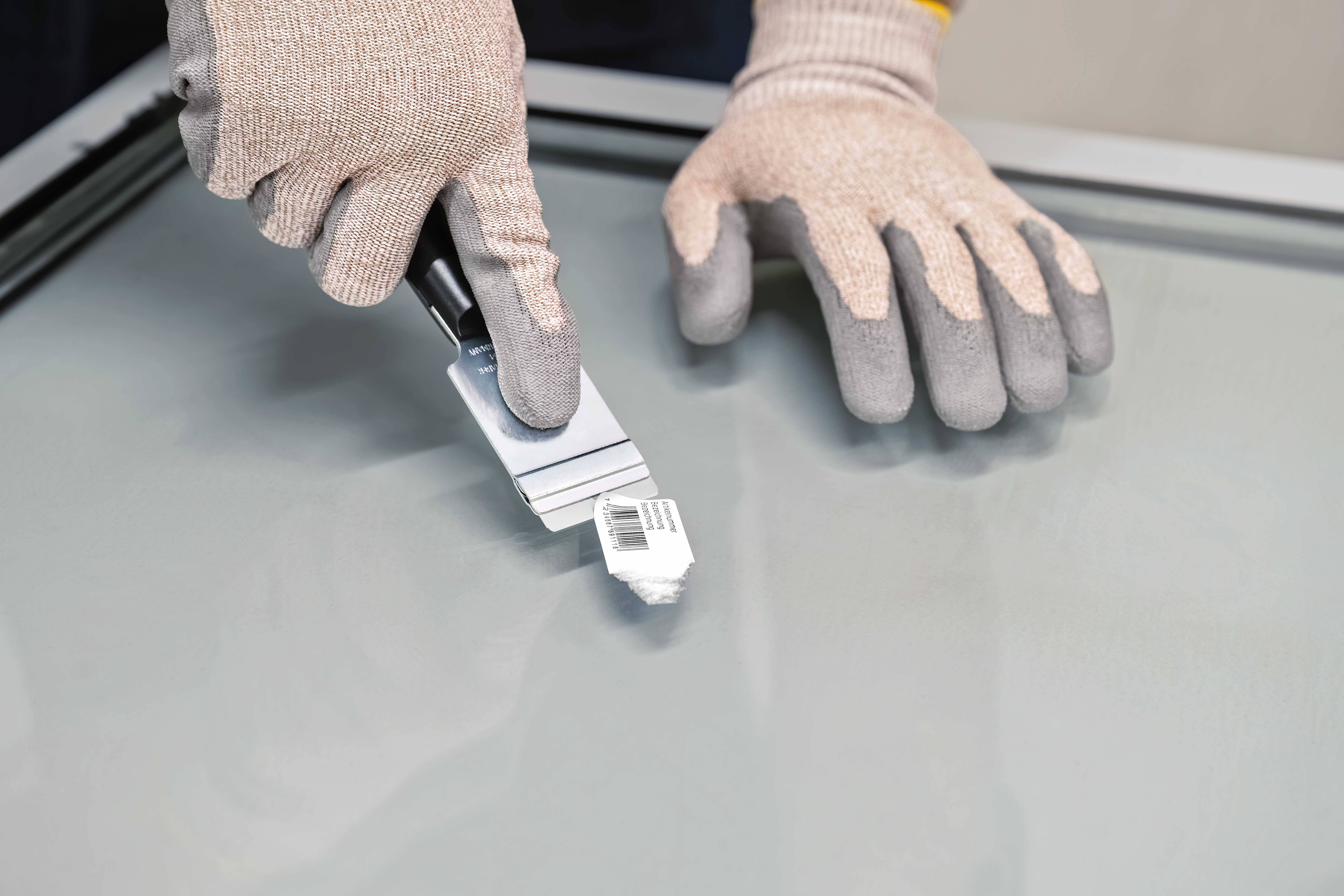 Une personne retire un autocollant d'une surface de verre lisse à l'aide d'un grattoir, tout en portant des gants de protection.