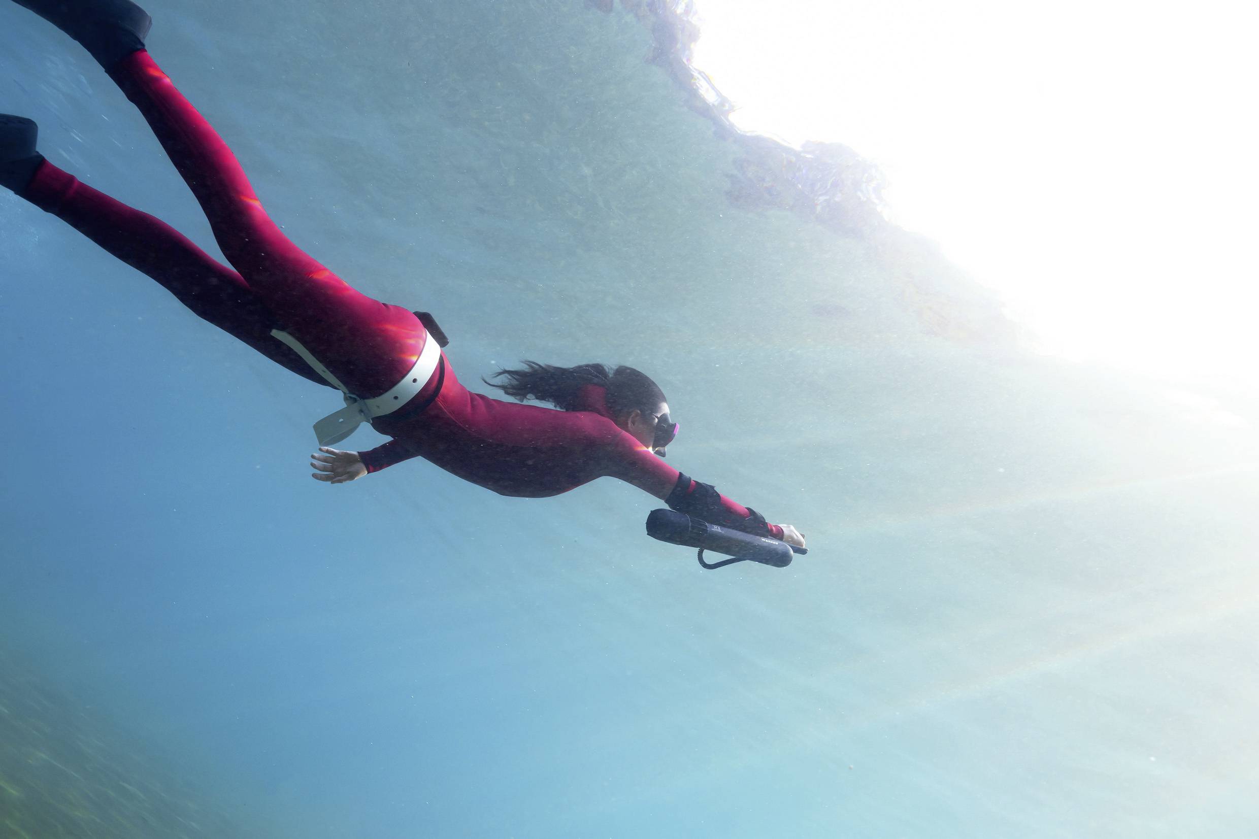 Une personne portant une combinaison de plongée rouge nage sous l'eau avec un appareil photo, entourée d'eau bleue et de lumière solaire venant d'en haut.