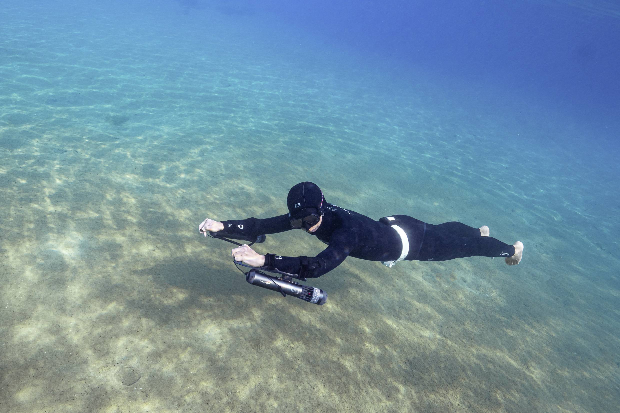 Un plongeur nage sous l'eau au-dessus d'un fond marin sablonneux, propulsé par un scooter sous-marin à main.