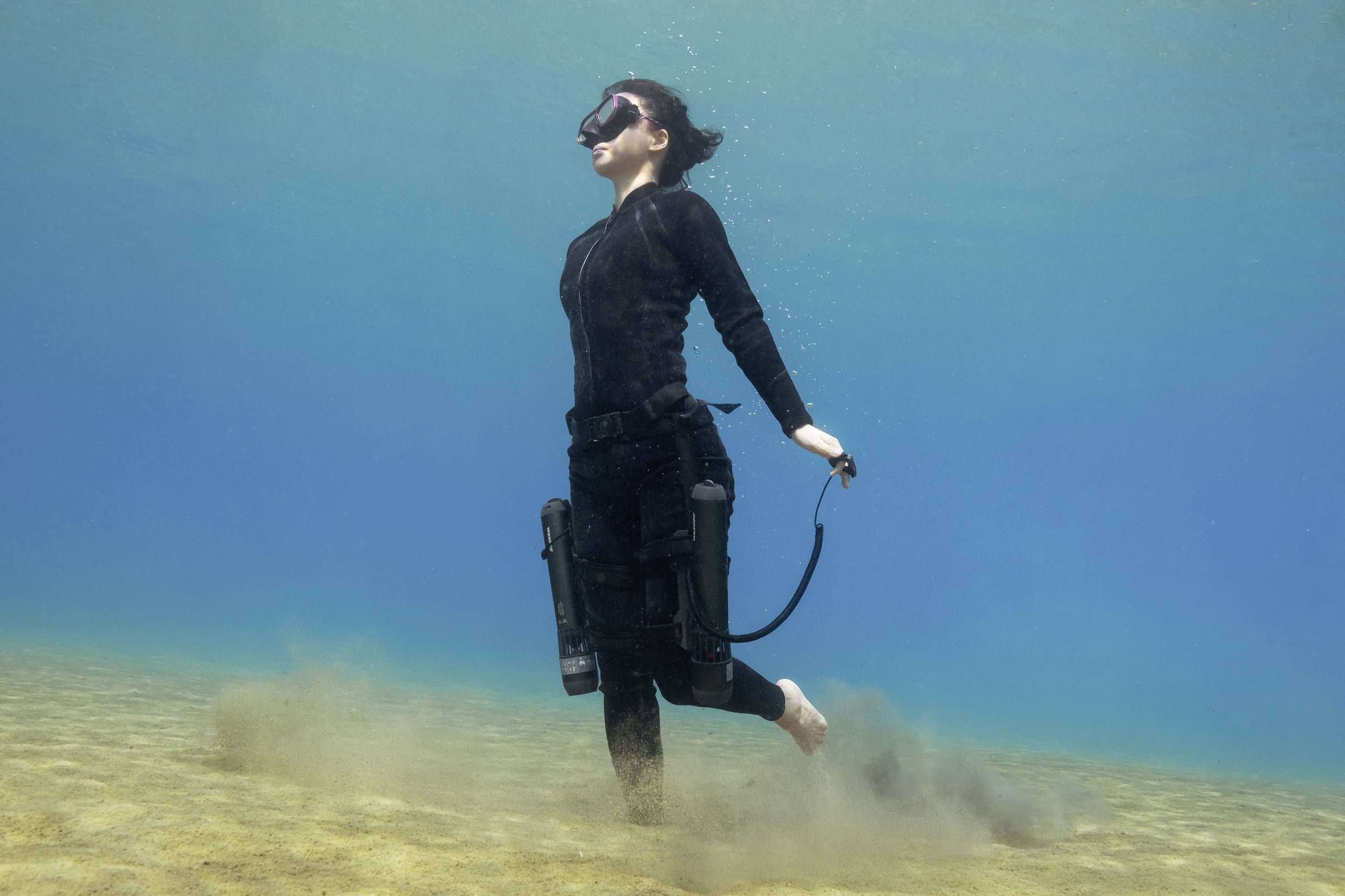 Une personne en combinaison de plongée noire flotte dans l'eau bleue et limpide, près du fond sablonneux, entourée de sable soulevé.
