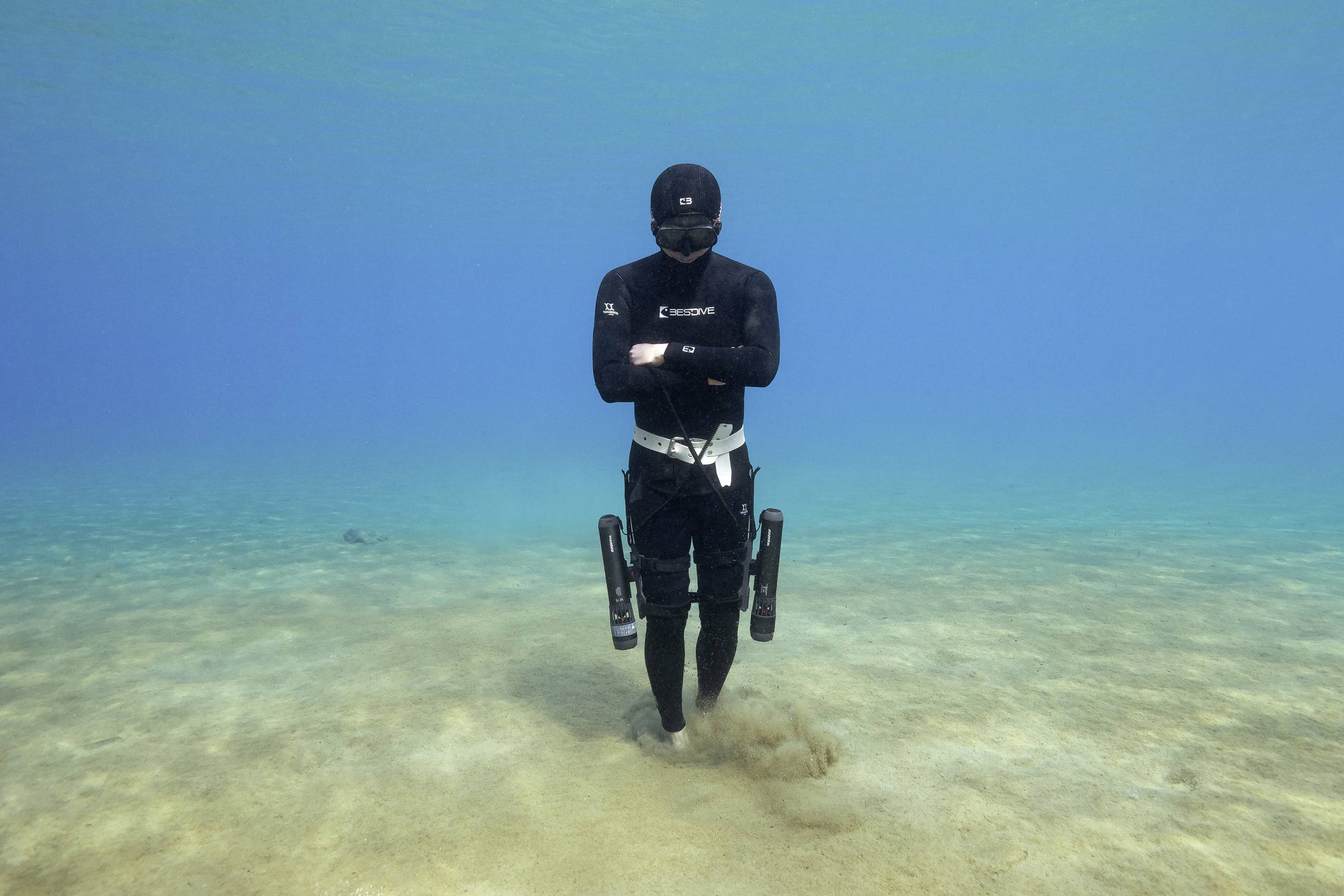 Un plongeur en combinaison de plongée noire marche sur le fond marin, entouré d'une eau cristalline.