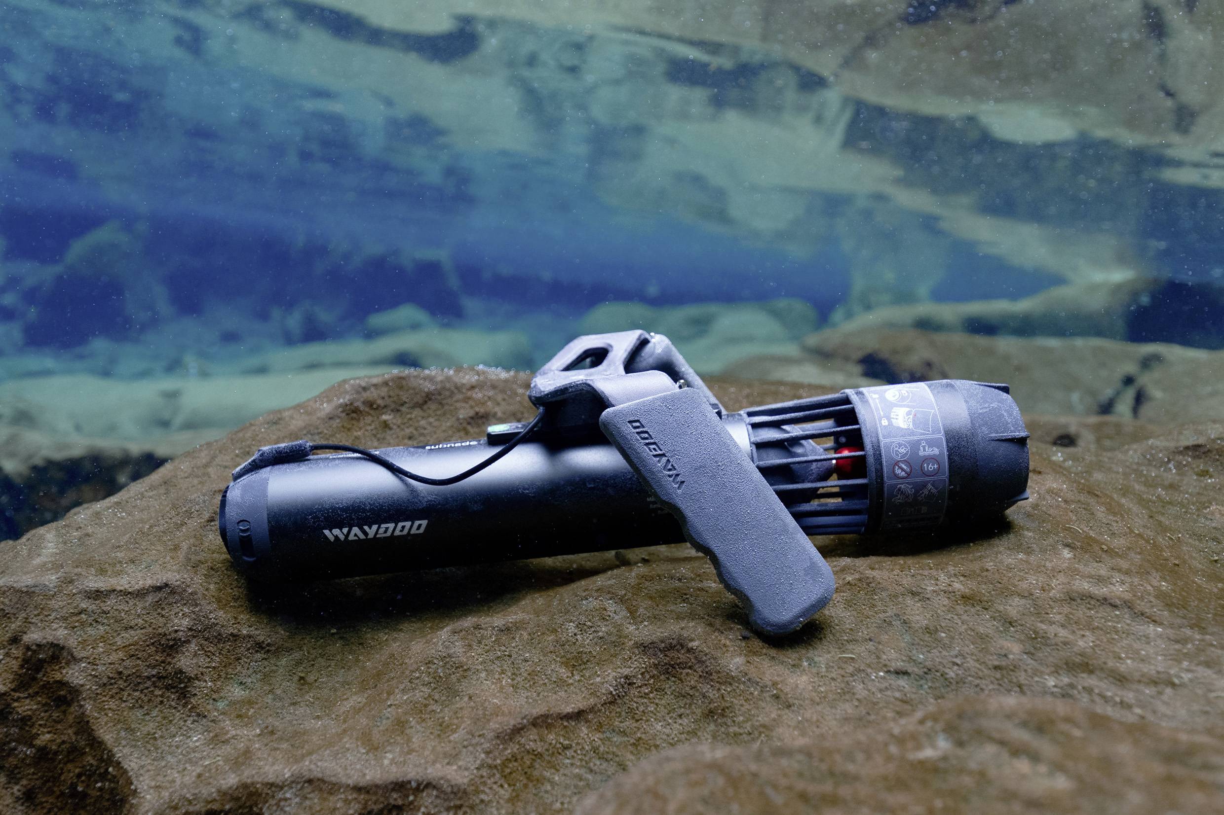 Un scooter sous-marin noir est posé sur un rocher sous l'eau. À l'arrière-plan, l'eau bleue est clairement visible.