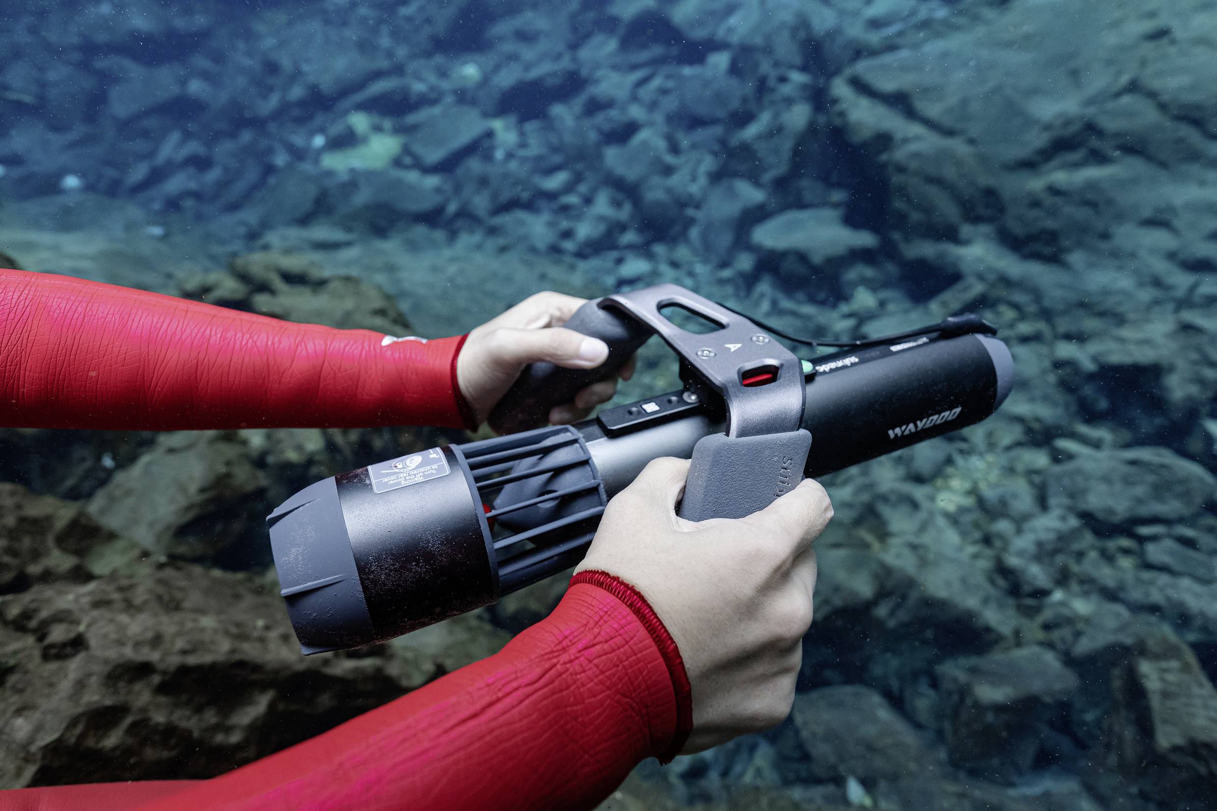 Une personne portant une combinaison de plongée rouge tient un scooter sous-marin dans un plan d'eau transparent, entourée de pierres.