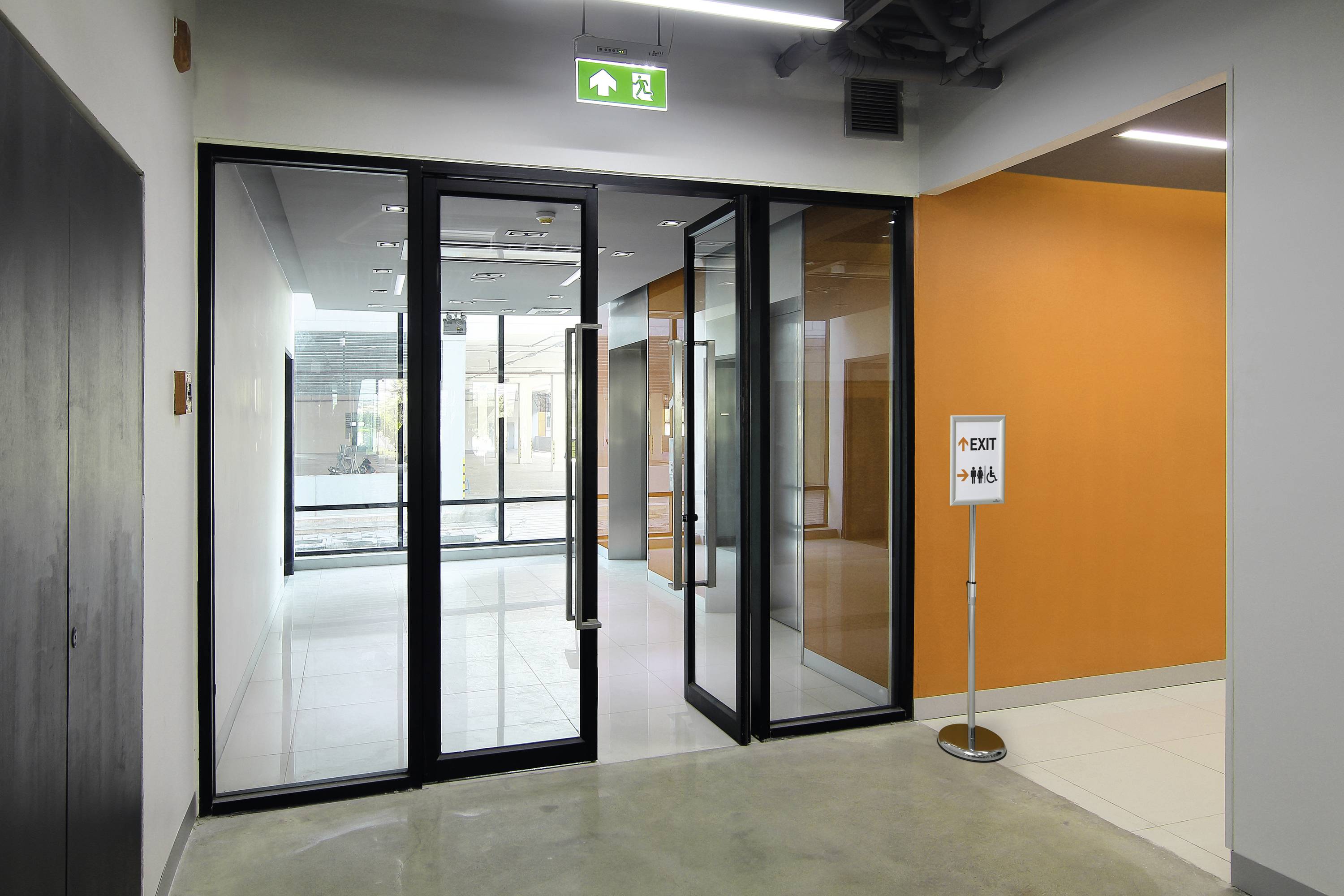 Portes vitrées doubles dans un couloir lumineux. Un panneau de sortie vert indique la voie de secours. Un panneau au sol signale « Sortie - H/F ».