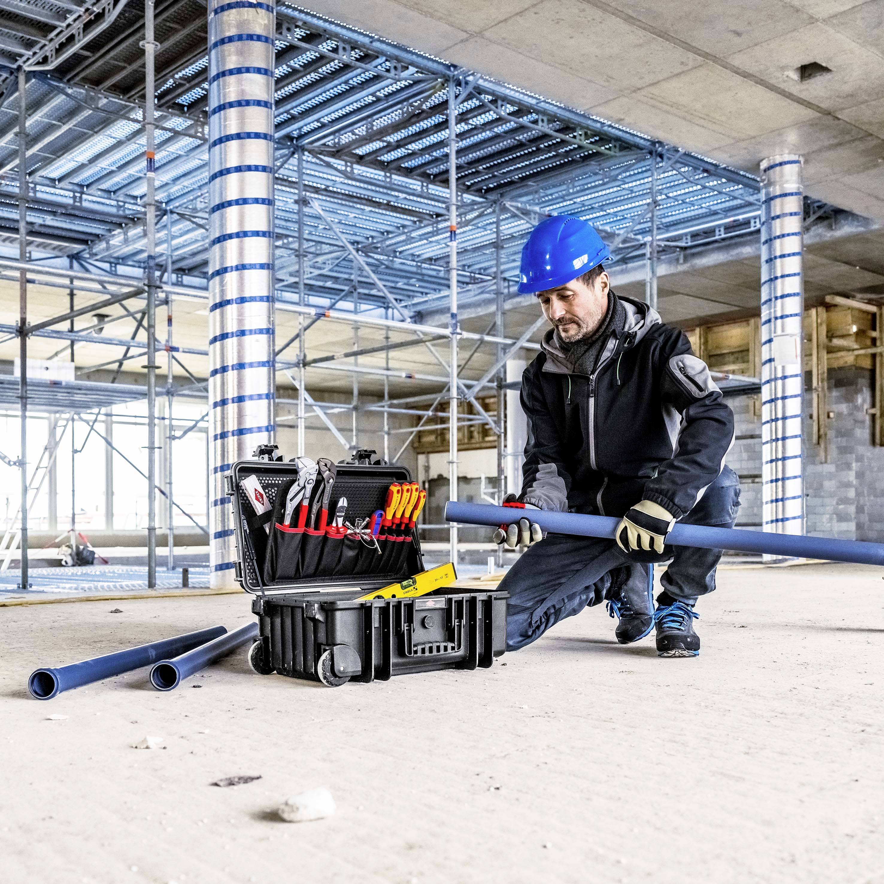Un ouvrier du bâtiment portant un casque de protection bleu est agenouillé sur un chantier, utilisant des outils d'une boîte à outils ouverte. Des échafaudages sont visibles en arrière-plan.