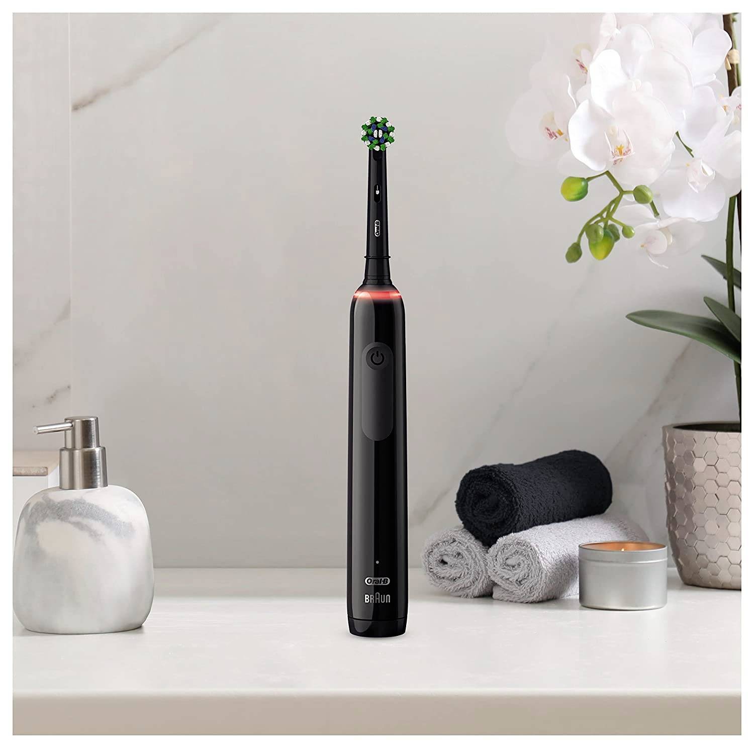 Une brosse à dents électrique est posée sur une étagère de salle de bain à côté d'une orchidée, de deux serviettes roulées et d'un distributeur de savon.