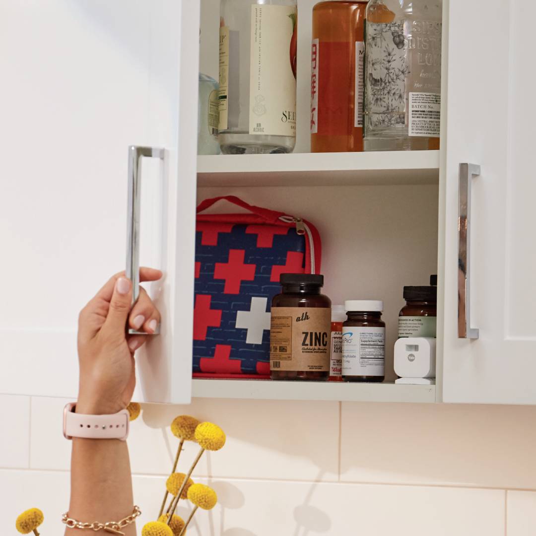 Une personne ouvre un placard de cuisine avec une main. À l'intérieur du placard sont visibles différentes bouteilles et un sac rouge avec une croix blanche.