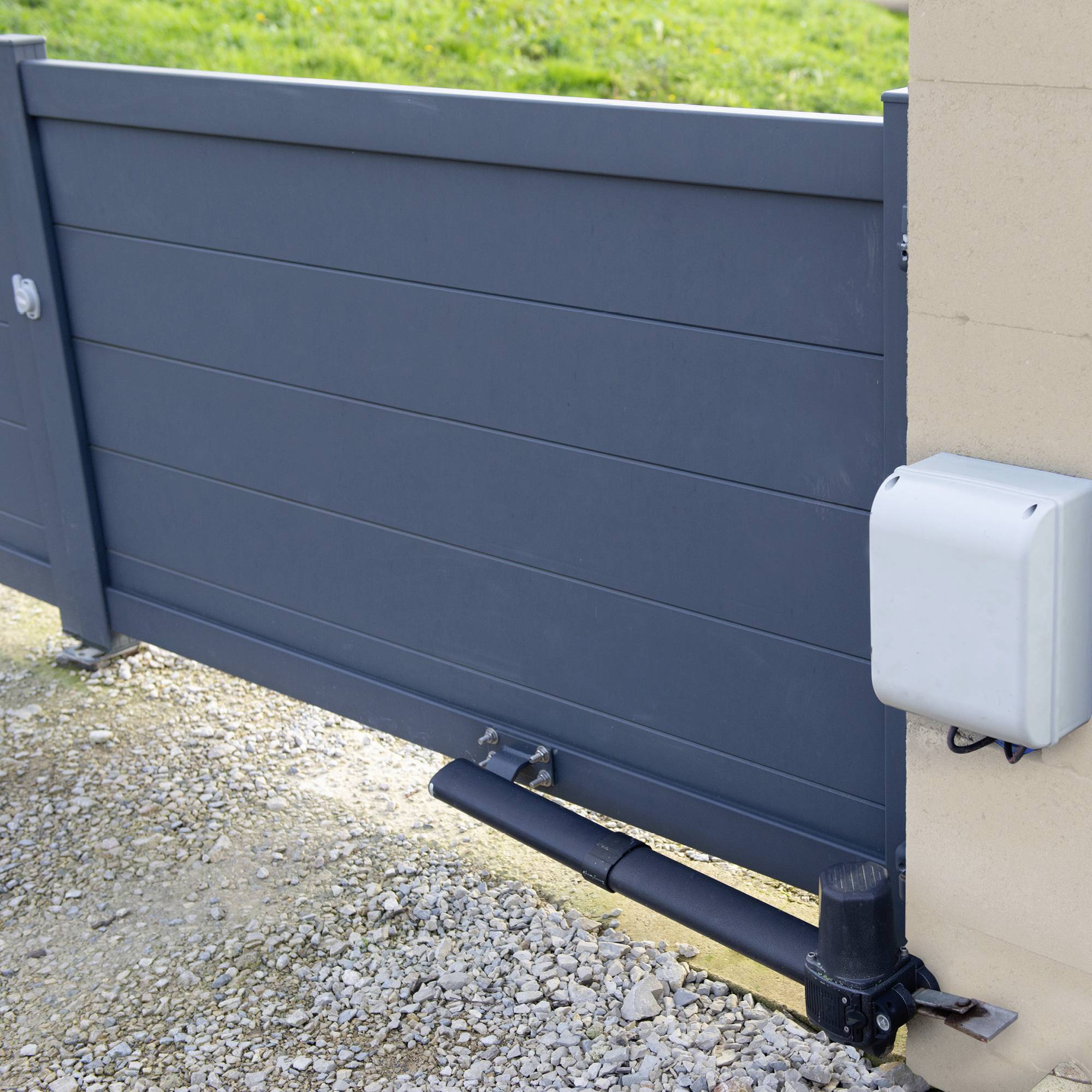 Porte métallique à entraînement et commande électriques, installée dans une allée de gravier, entourée de surfaces enherbées.