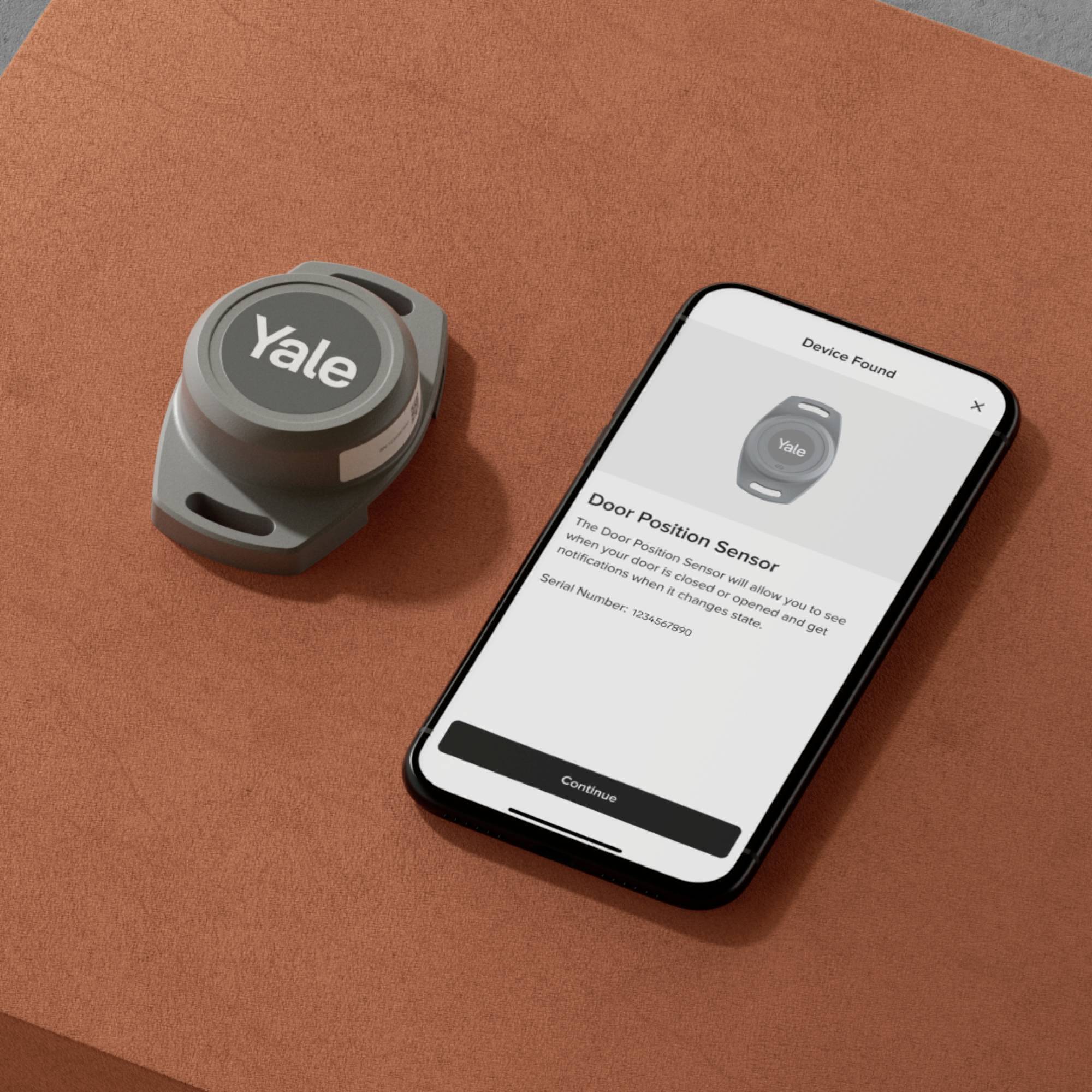 Un capteur de position de porte gris à côté d'un smartphone sur une surface orange. L'écran affiche la connexion au capteur.