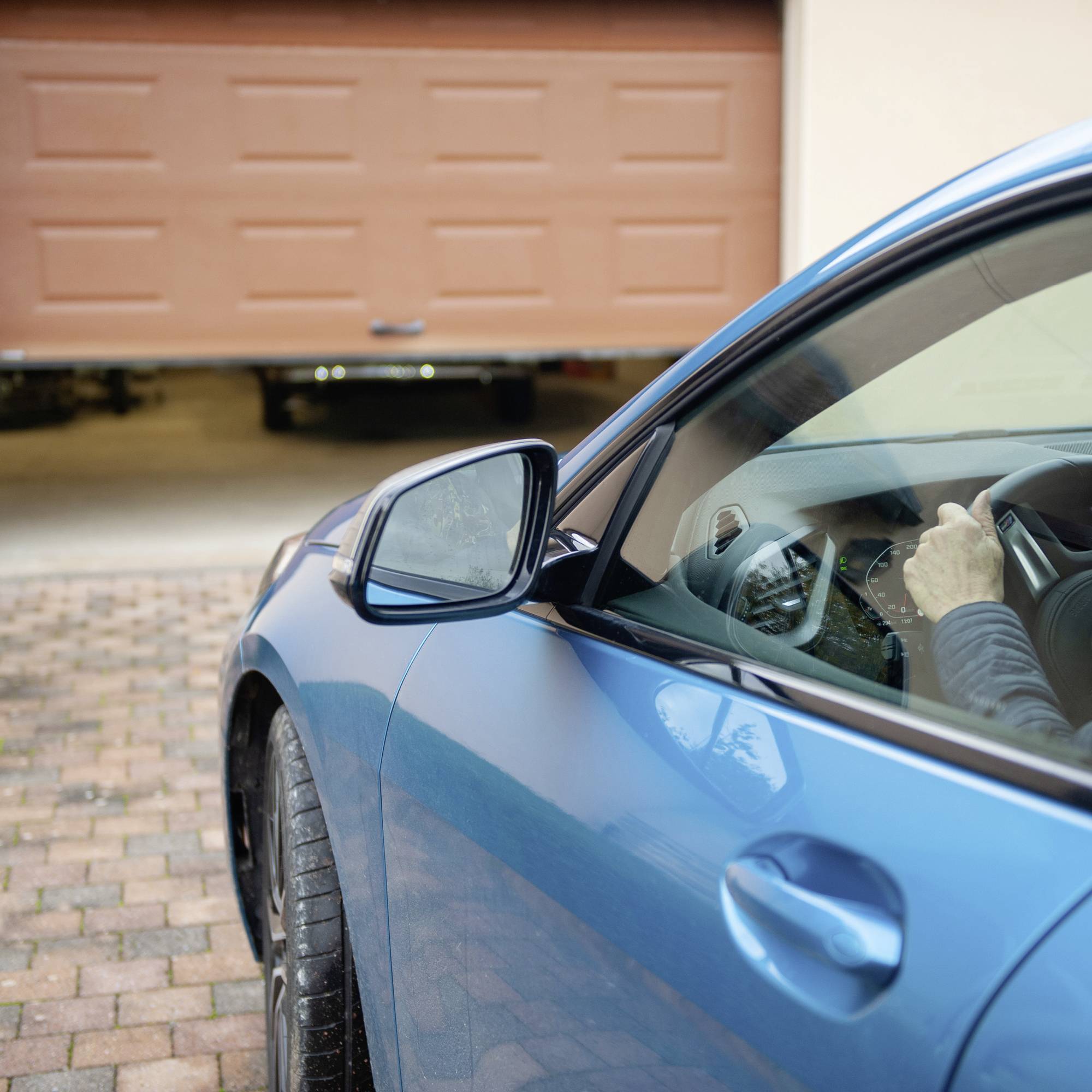 'Gros plan sur une voiture bleue entrant dans un garage ouvert. Mains du conducteur visibles sur le volant. Porte de garage partiellement ouverte.'