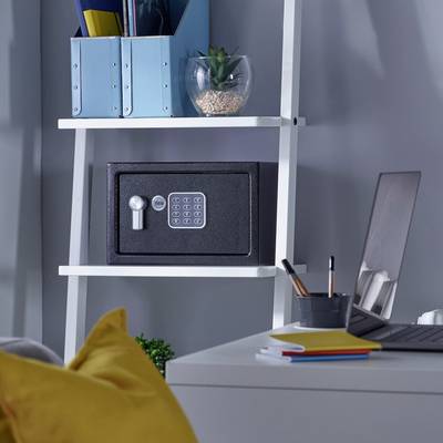 Une étagère blanche avec un coffre-fort, des livres et un verre contenant une plante posés dessus. Un ordinateur portable et un carnet de notes sont placés sur un bureau à côté.