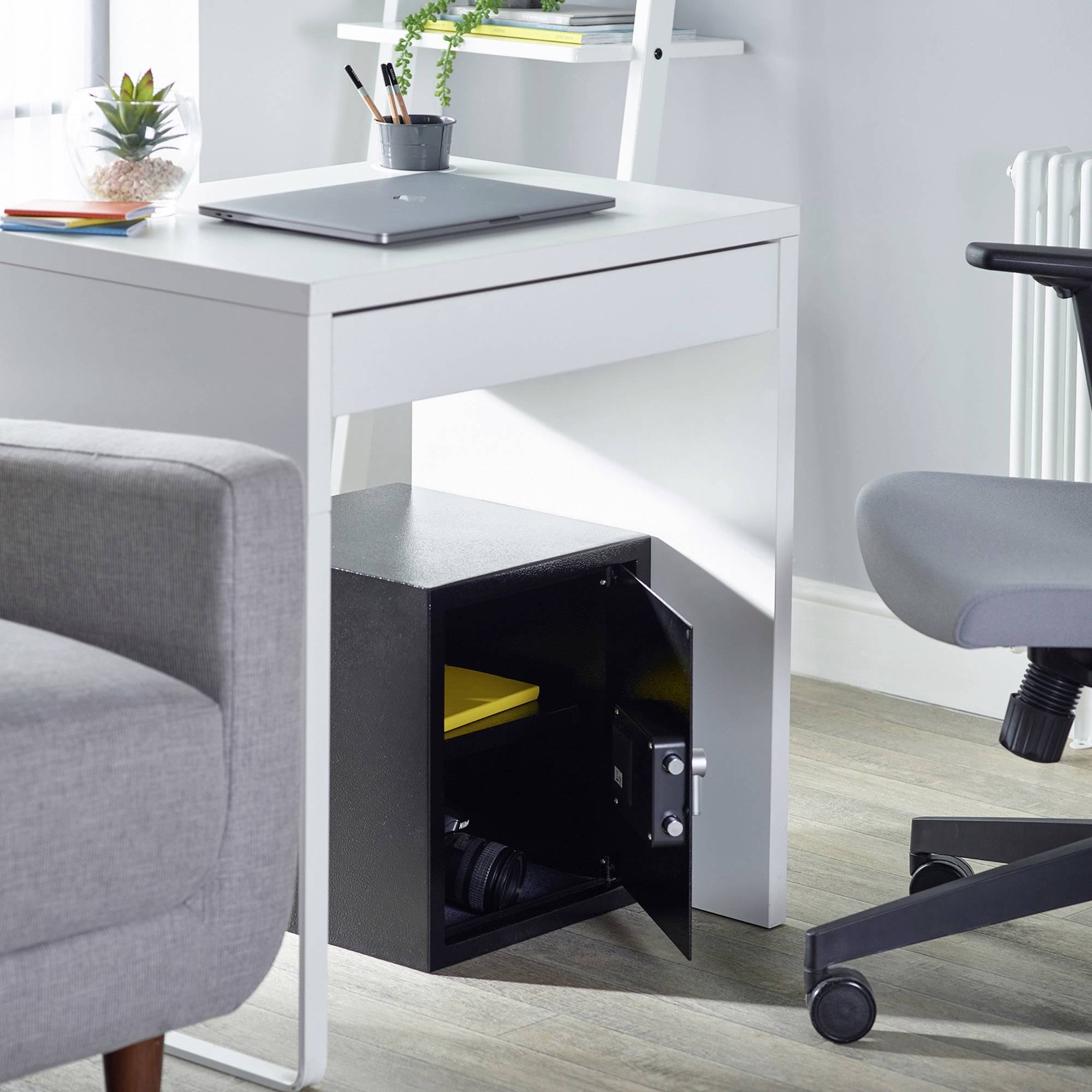 Un coffre-fort ouvert sous un bureau révèle des documents jaunes et un appareil photo à l'intérieur. Un ordinateur portable et des plantes sont posés sur le bureau.